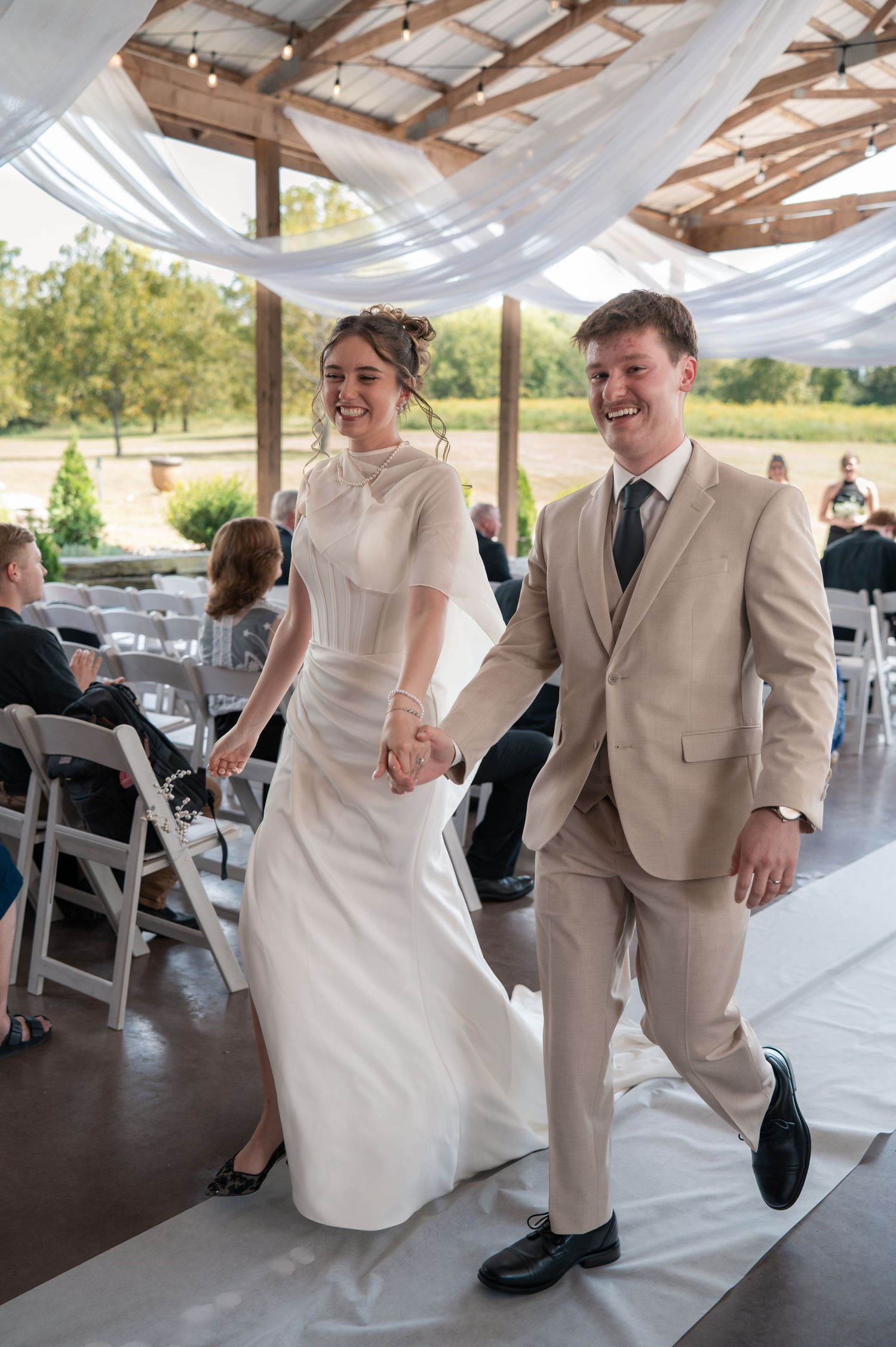 Joyful newlyweds walk hand-in-hand down the aisle at an outdoor barn wedding ceremony.