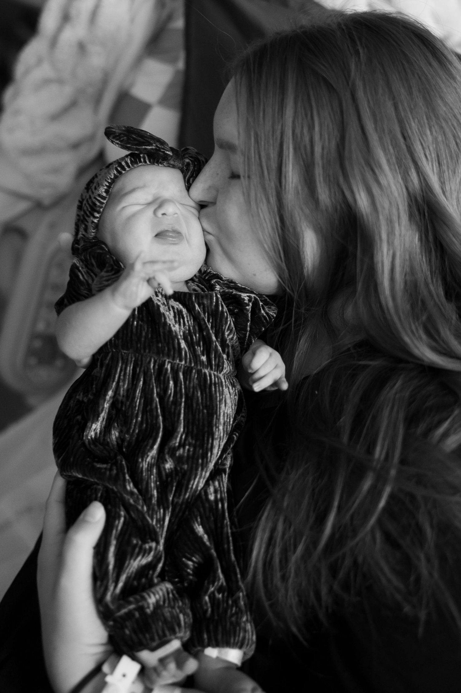 Black and white photo of a mother kissing her newborn baby dressed in a velvet floral outfit.