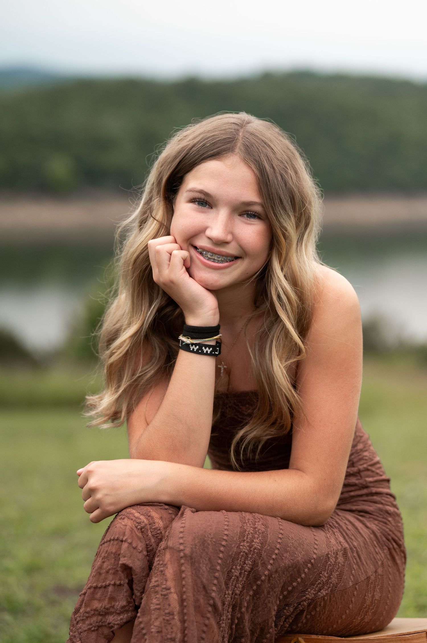 Smiling teenage girl with braces posing outdoors near a lake in a brown strapless outfit.