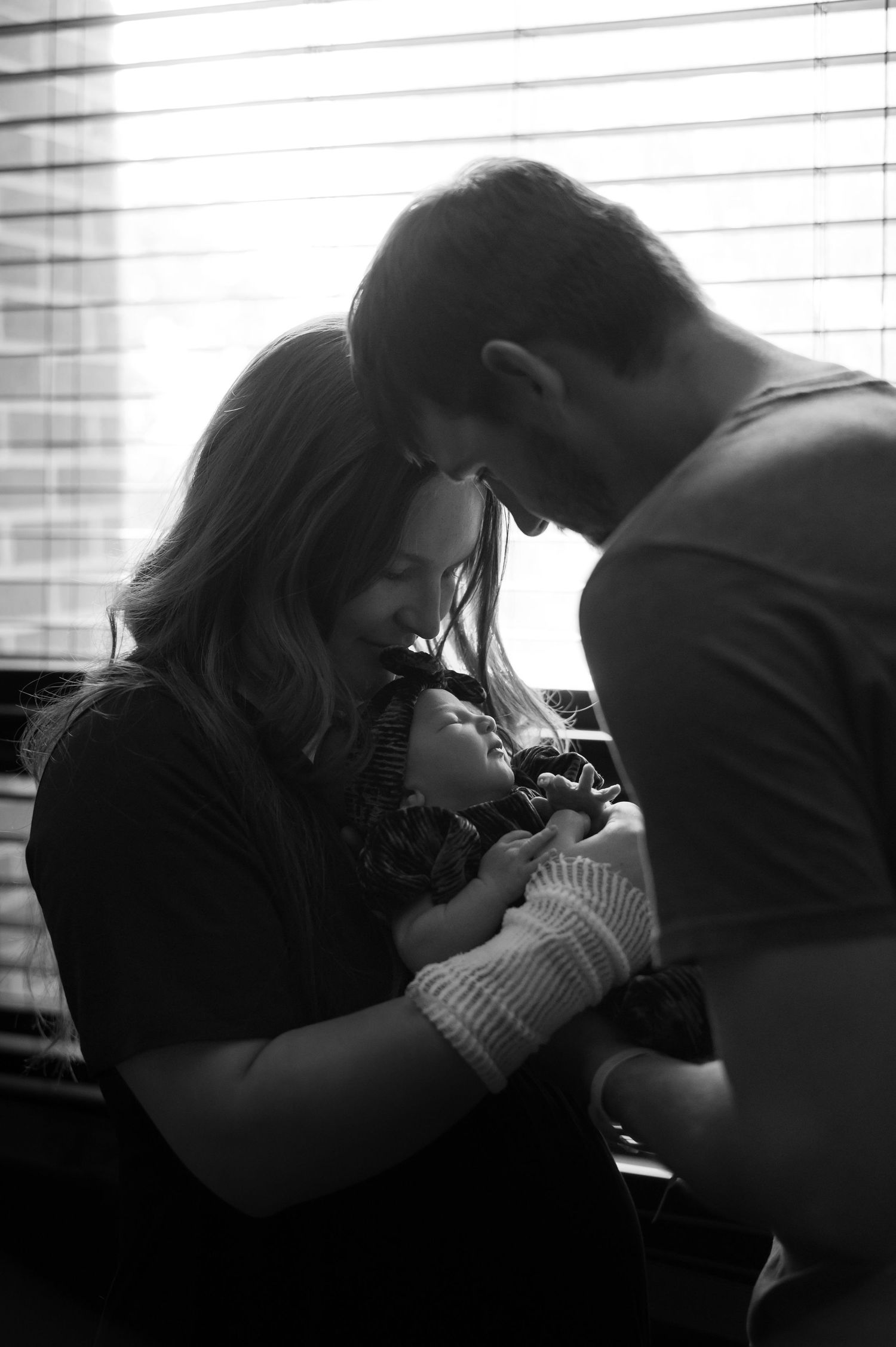 Black and white silhouette of new parents lovingly cradling their newborn baby near a window.