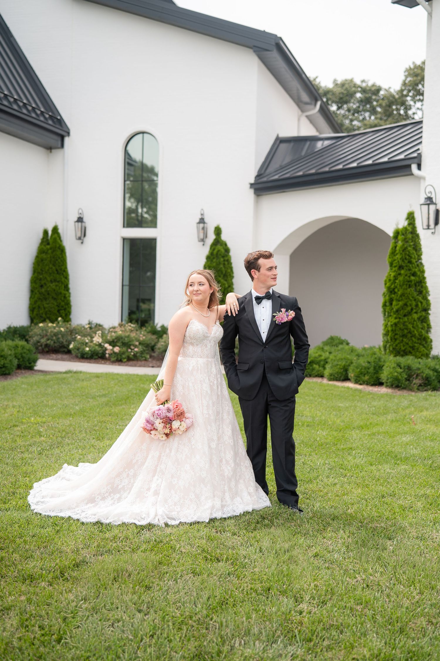 Bride in lace ball gown and groom in black tuxedo pose together on green lawn outside elegant white venue.