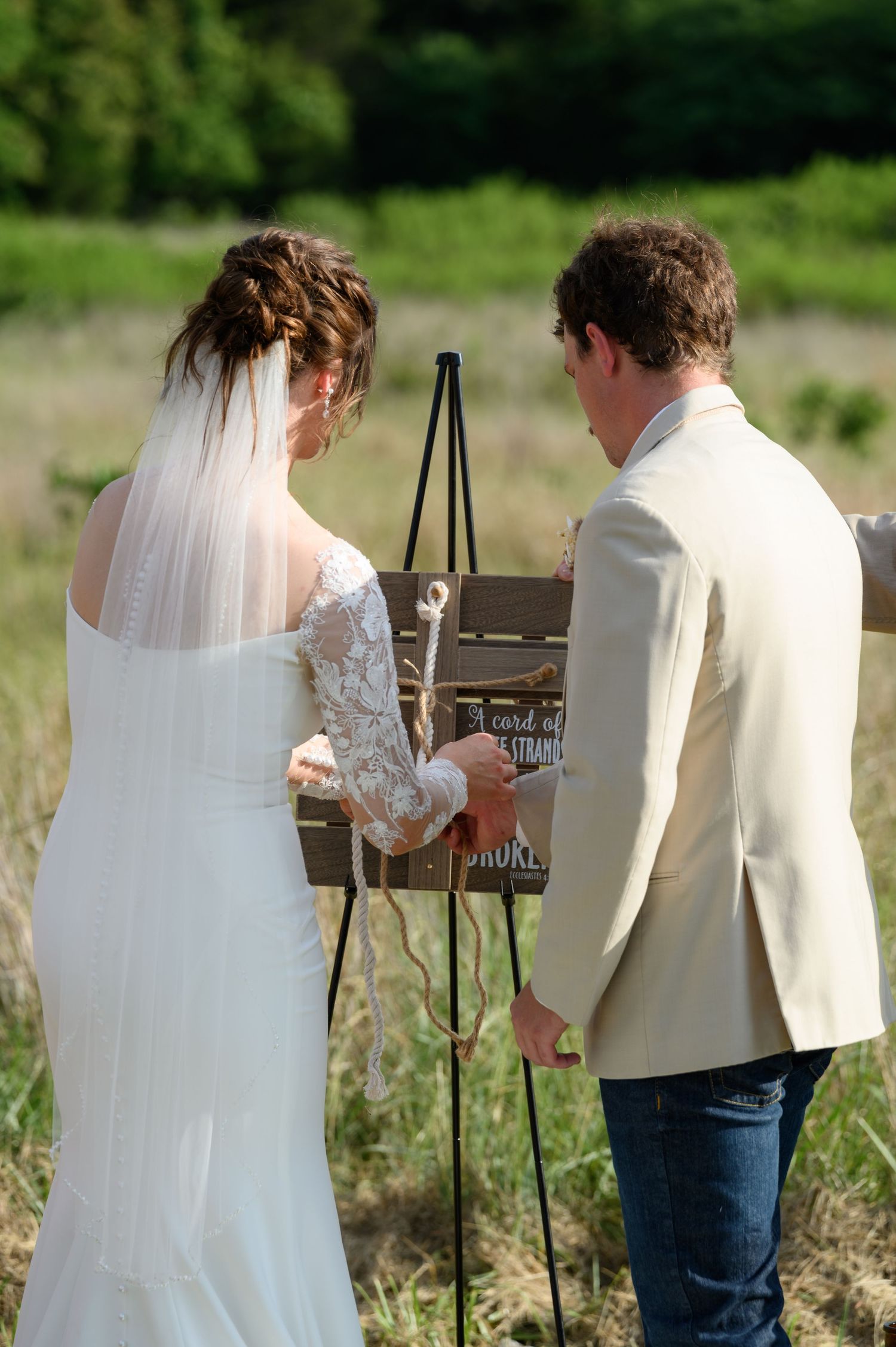 Bride and groom perform cord of three strands unity ritual at rustic outdoor wedding ceremony.