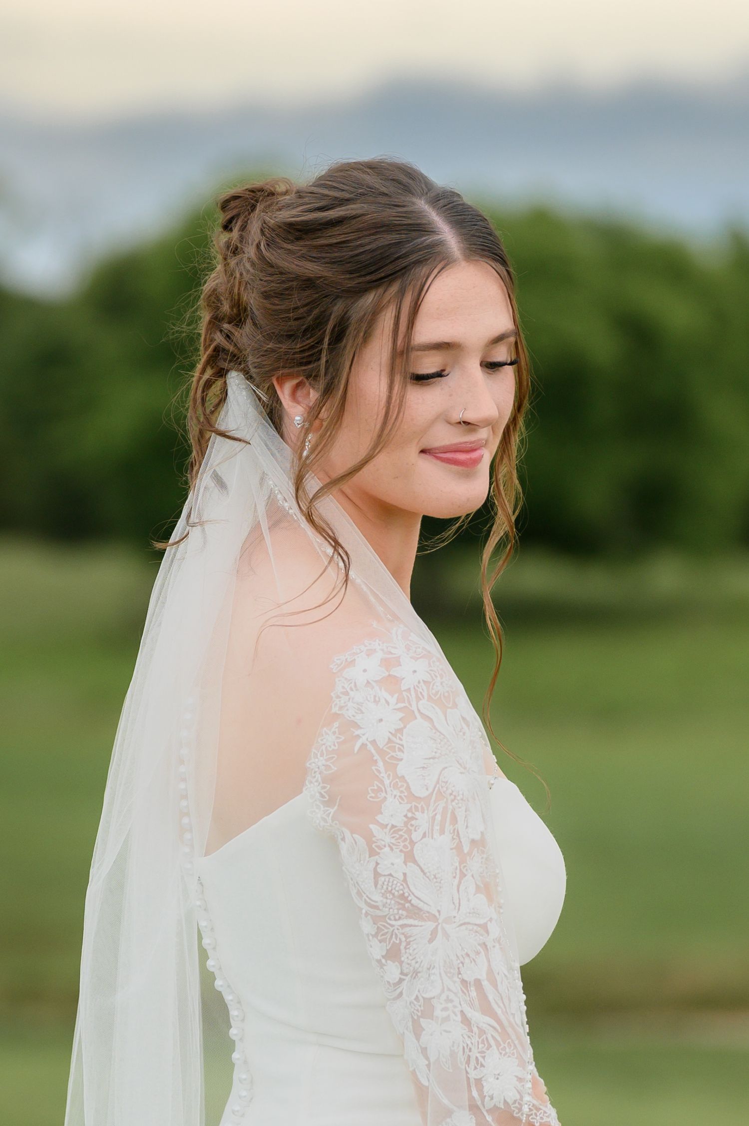 Elegant bride in lace wedding gown and veil with updo hairstyle posing outdoors at golden hour.
