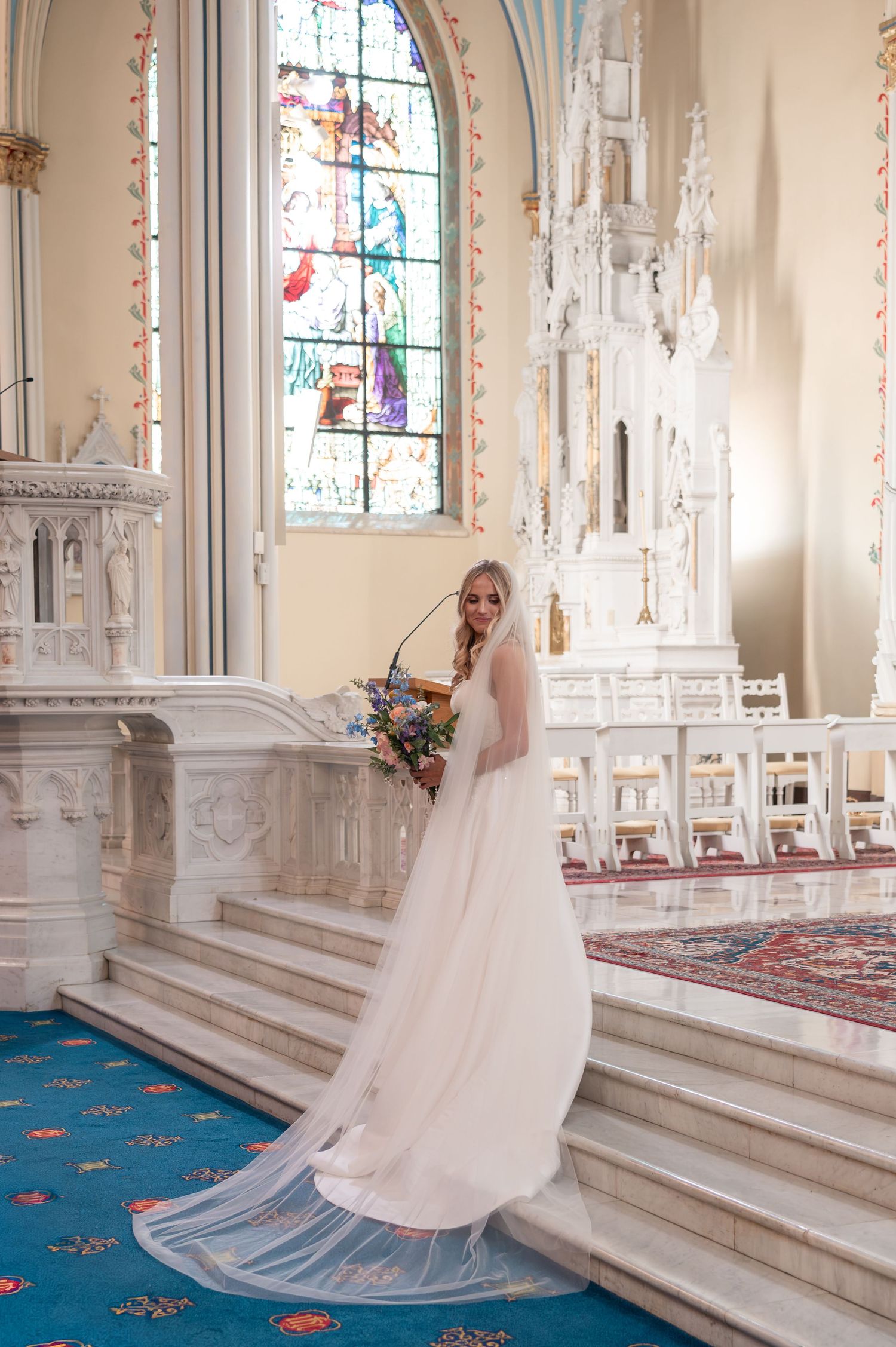 Elegant bride in flowing white gown poses on church altar steps beside colorful stained glass windows.
