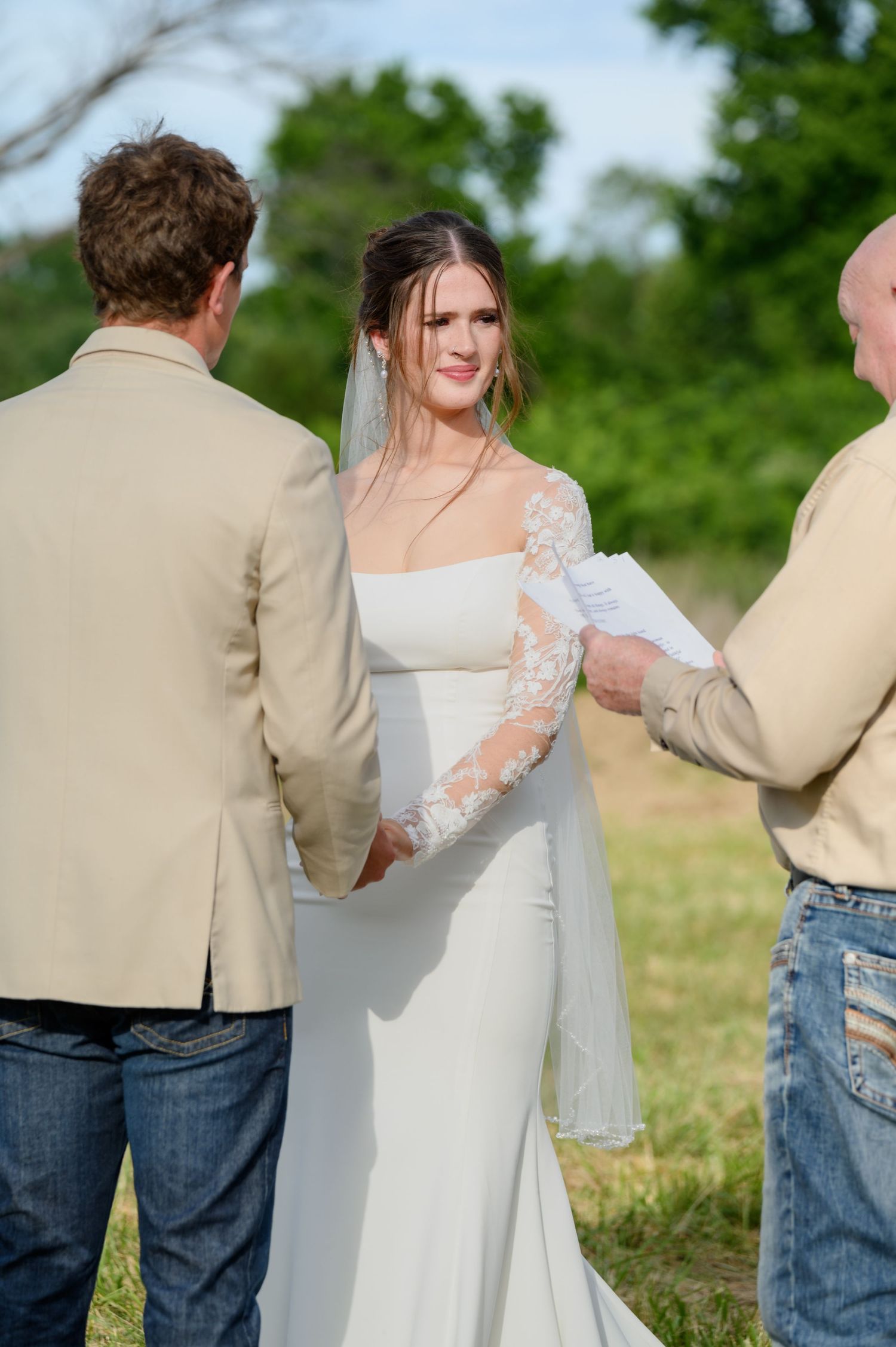 Bride in lace sleeve white gown exchanges vows with groom in tan suit at outdoor ceremony.
