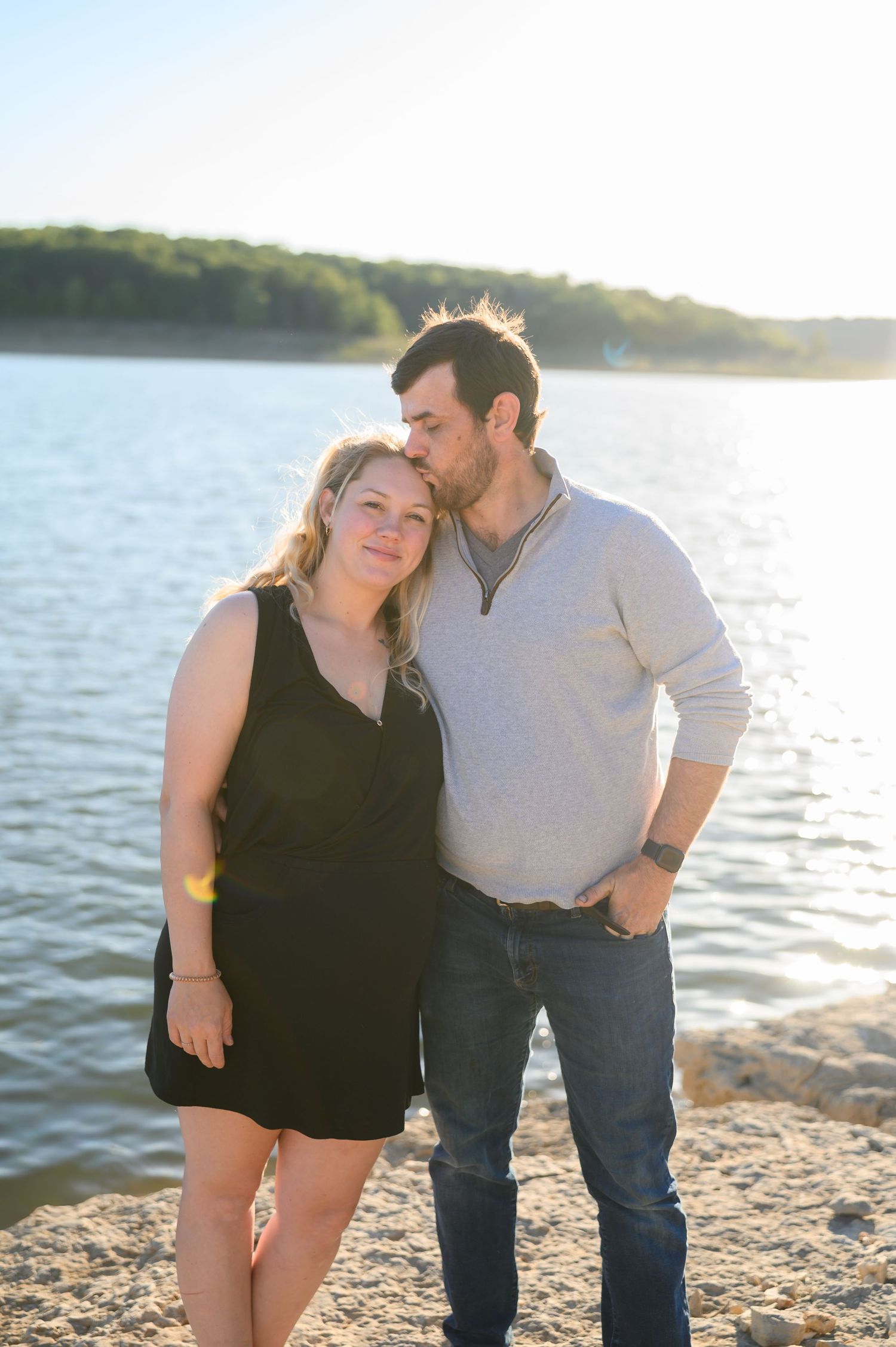 Man kissing woman on the head as they pose together on a sunny rocky lakeside.