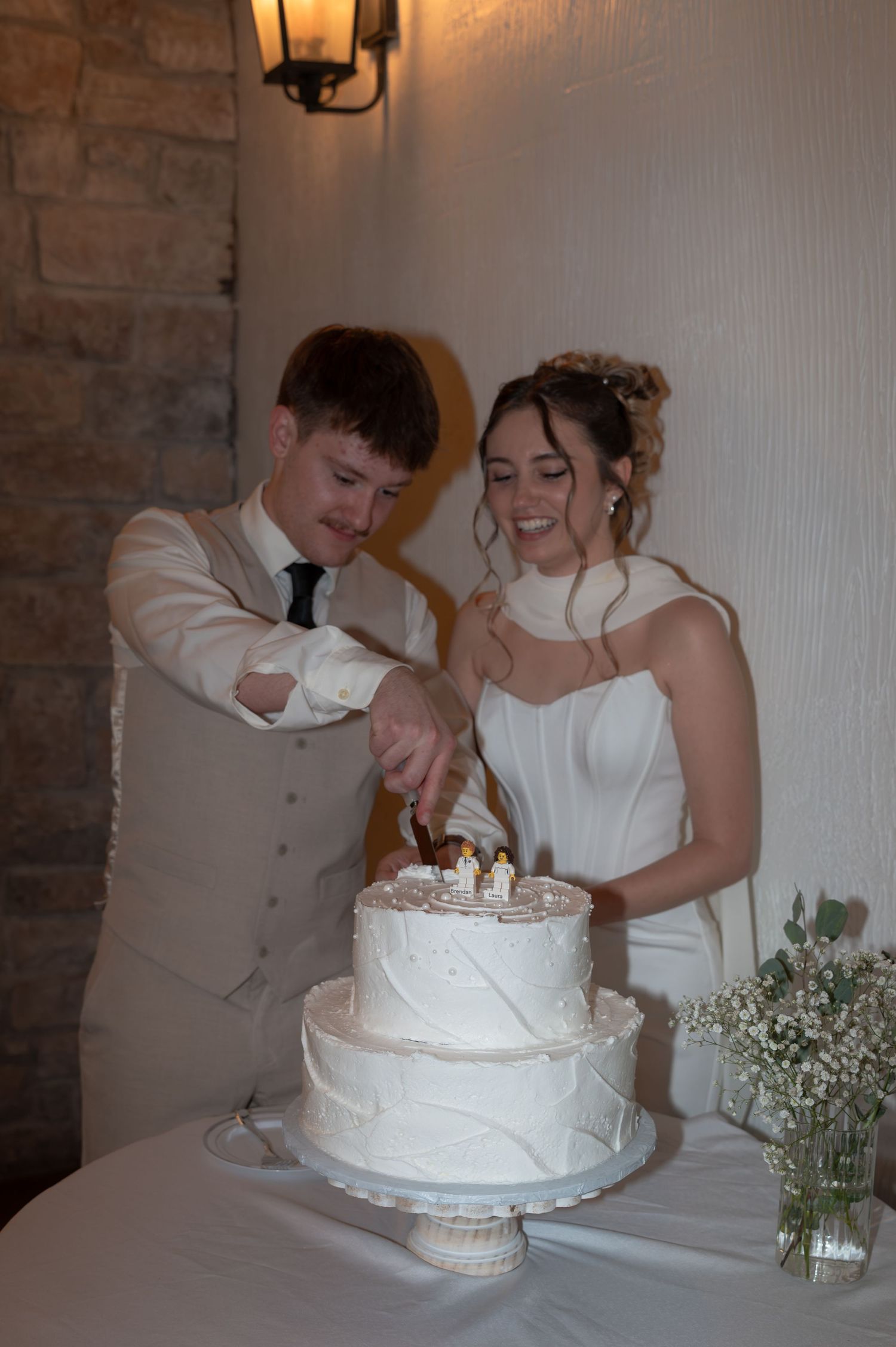 Bride and groom cutting a two-tier white wedding cake together, smiling, with floral decor nearby.