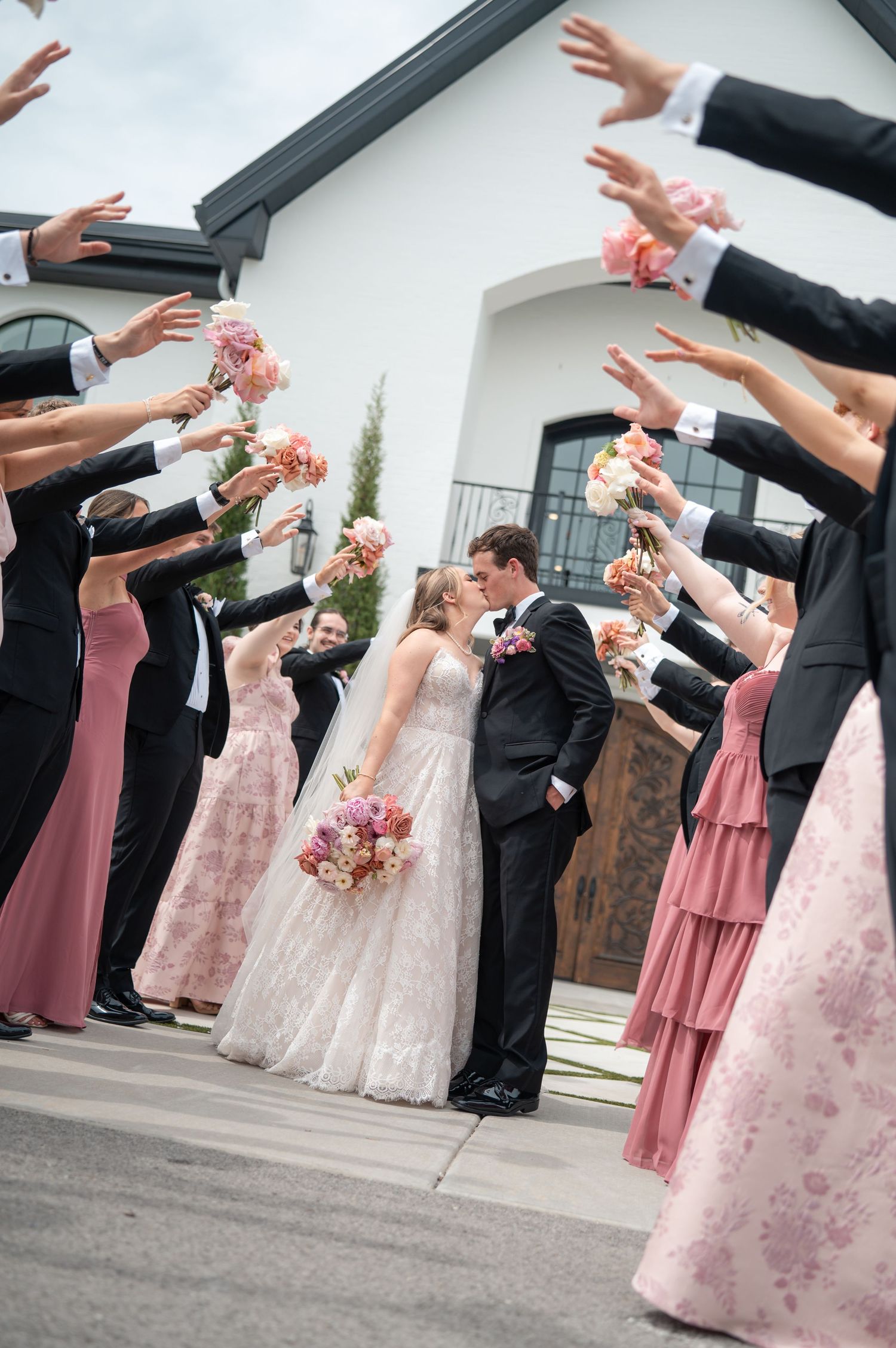 Wedding party tunnel kiss with bridesmaids in pink dresses and groomsmen in black tuxedos outside white chapel.