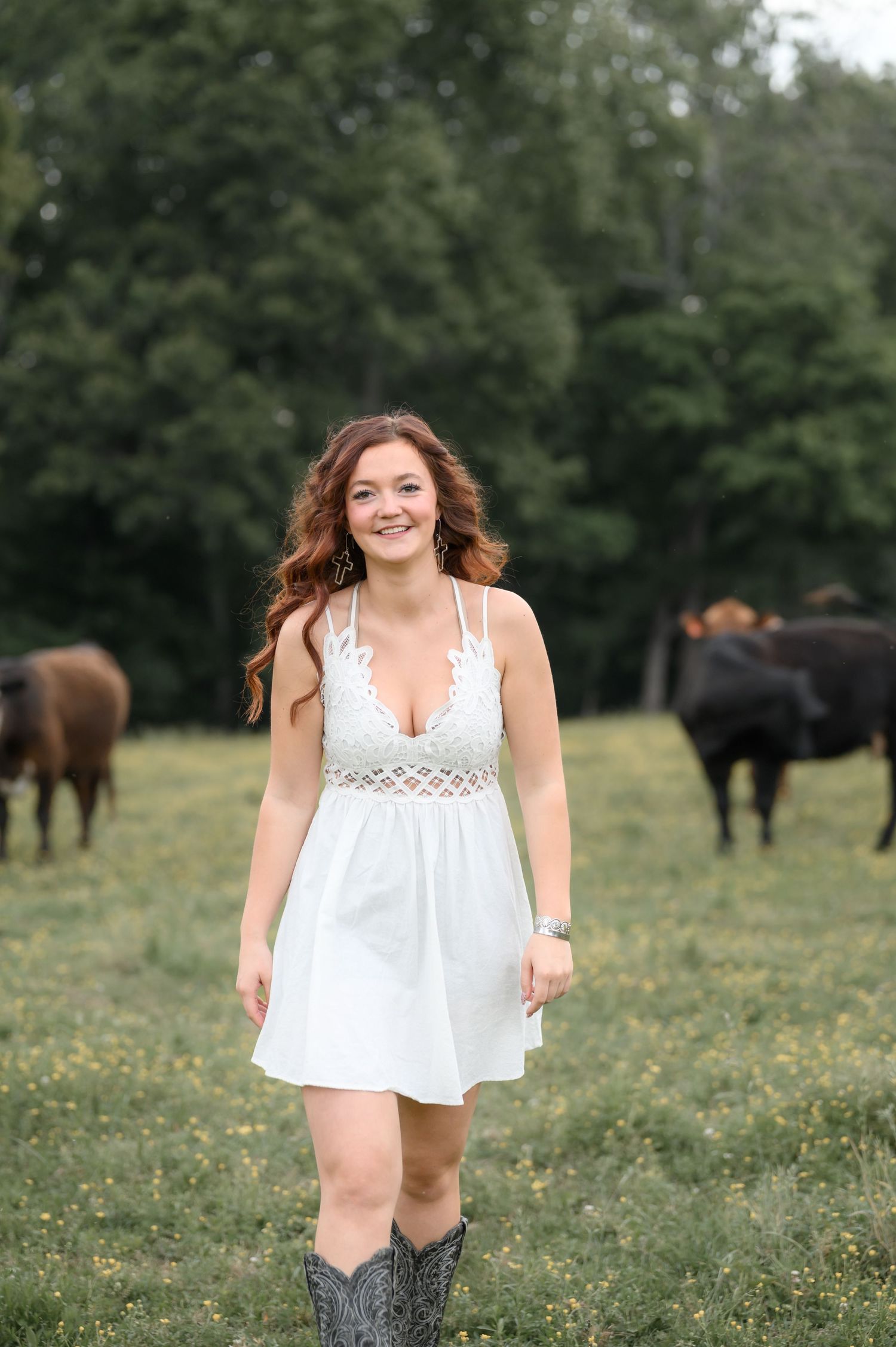 Young woman in white crochet dress and cowboy boots smiling in a green pasture with cattle.