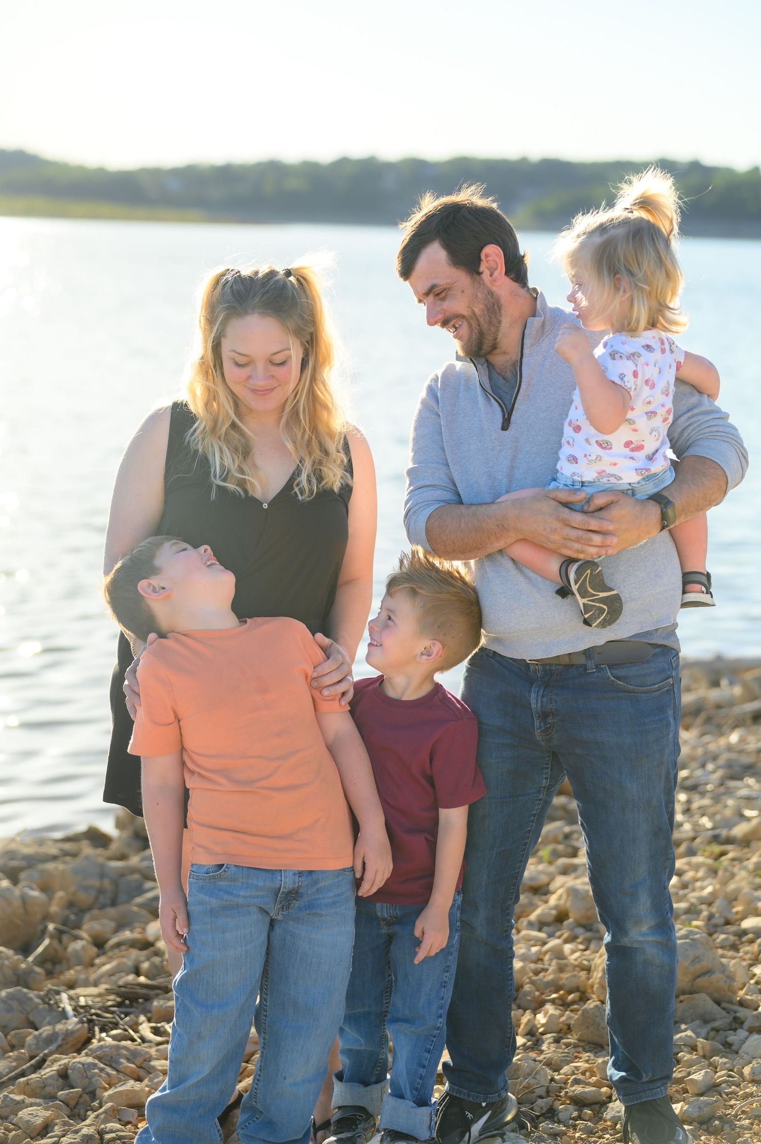 Happy family of five laughing together on rocky lakeshore in warm golden sunset light.