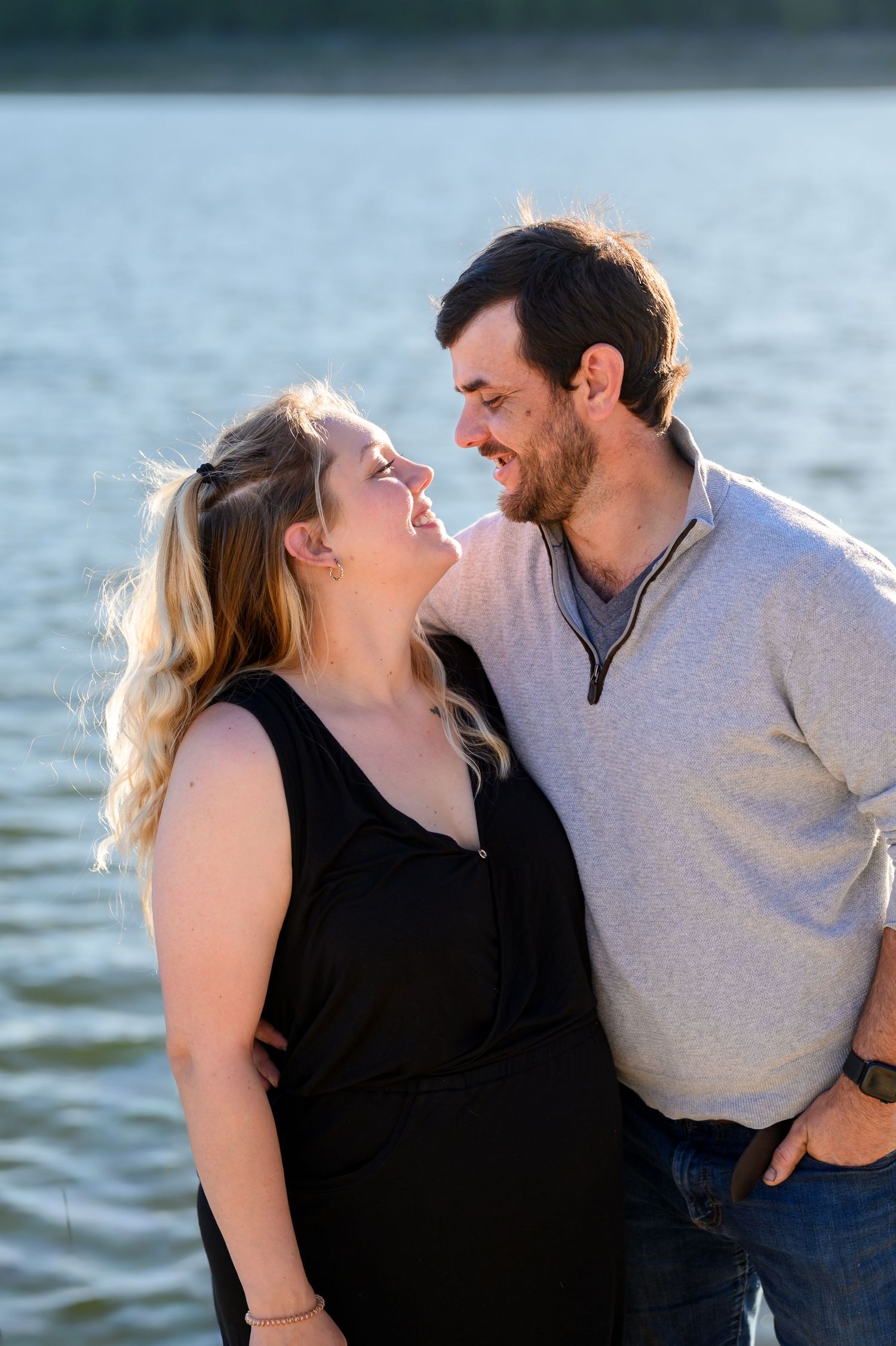 Couple smiling at each other standing close together in front of a sunlit lake at golden hour.