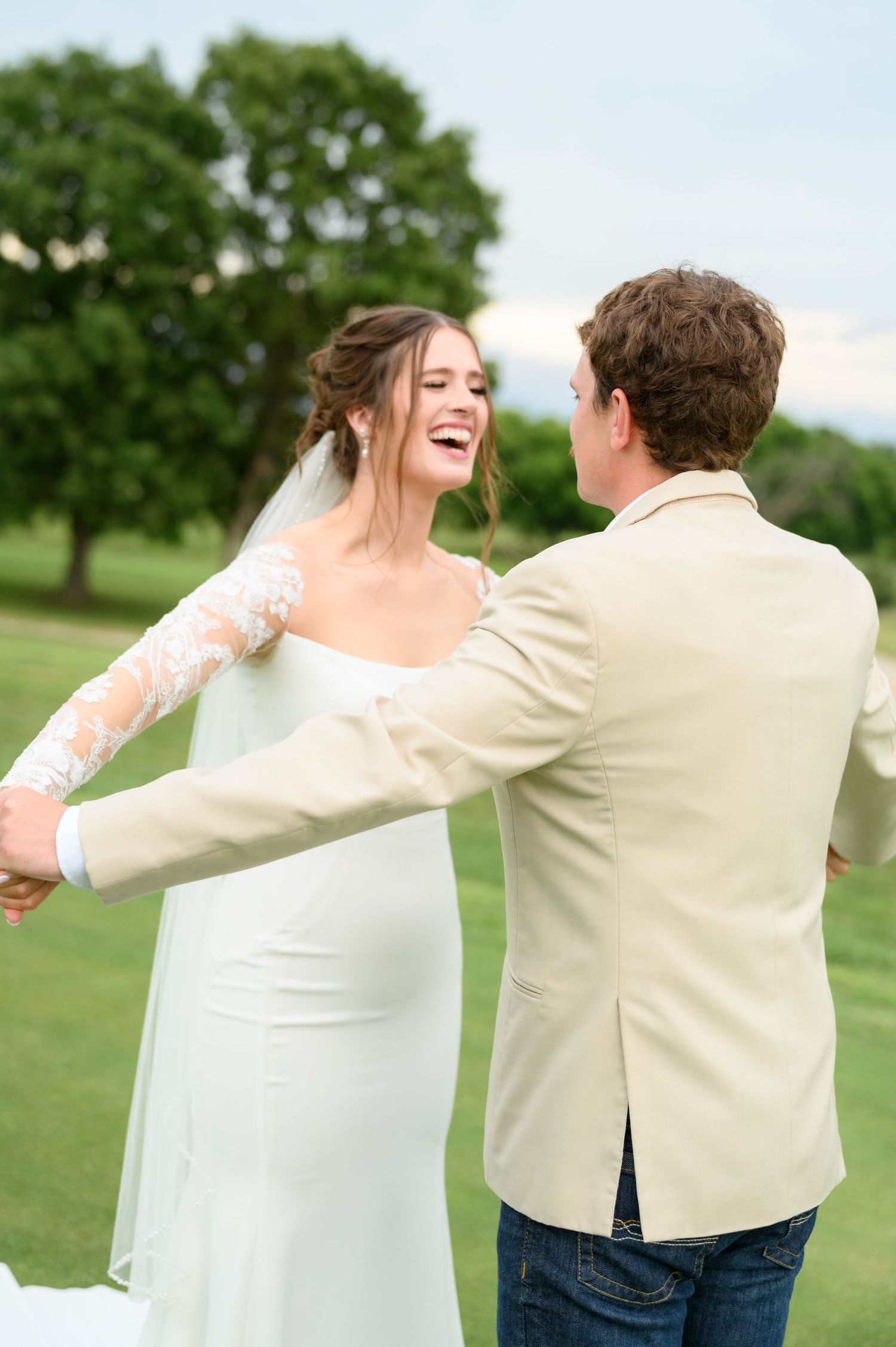 Bride in white lace wedding dress laughing joyfully while dancing with groom in tan blazer outdoors.