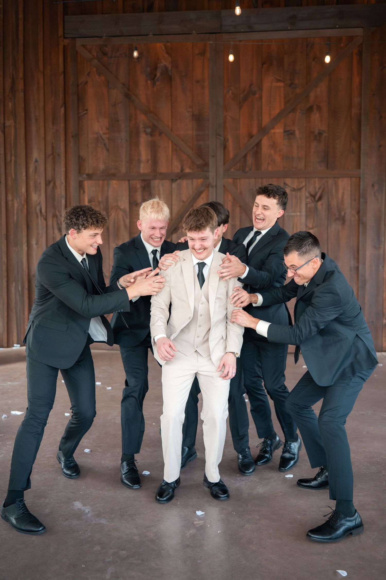 Groom in ivory suit laughing with four groomsmen in black suits outside rustic barn doors.