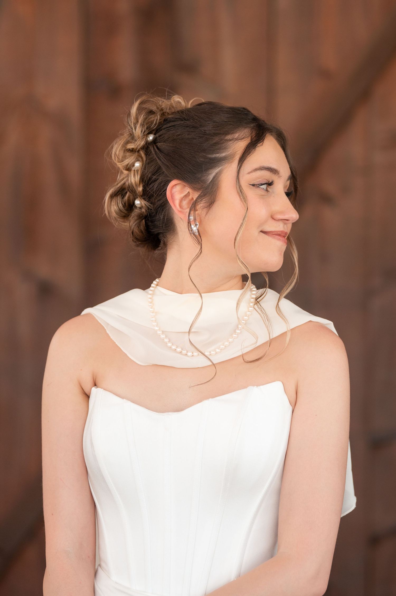 Young bride in white gown with pearl necklace and updo hairstyle smiling softly against rustic wood backdrop.