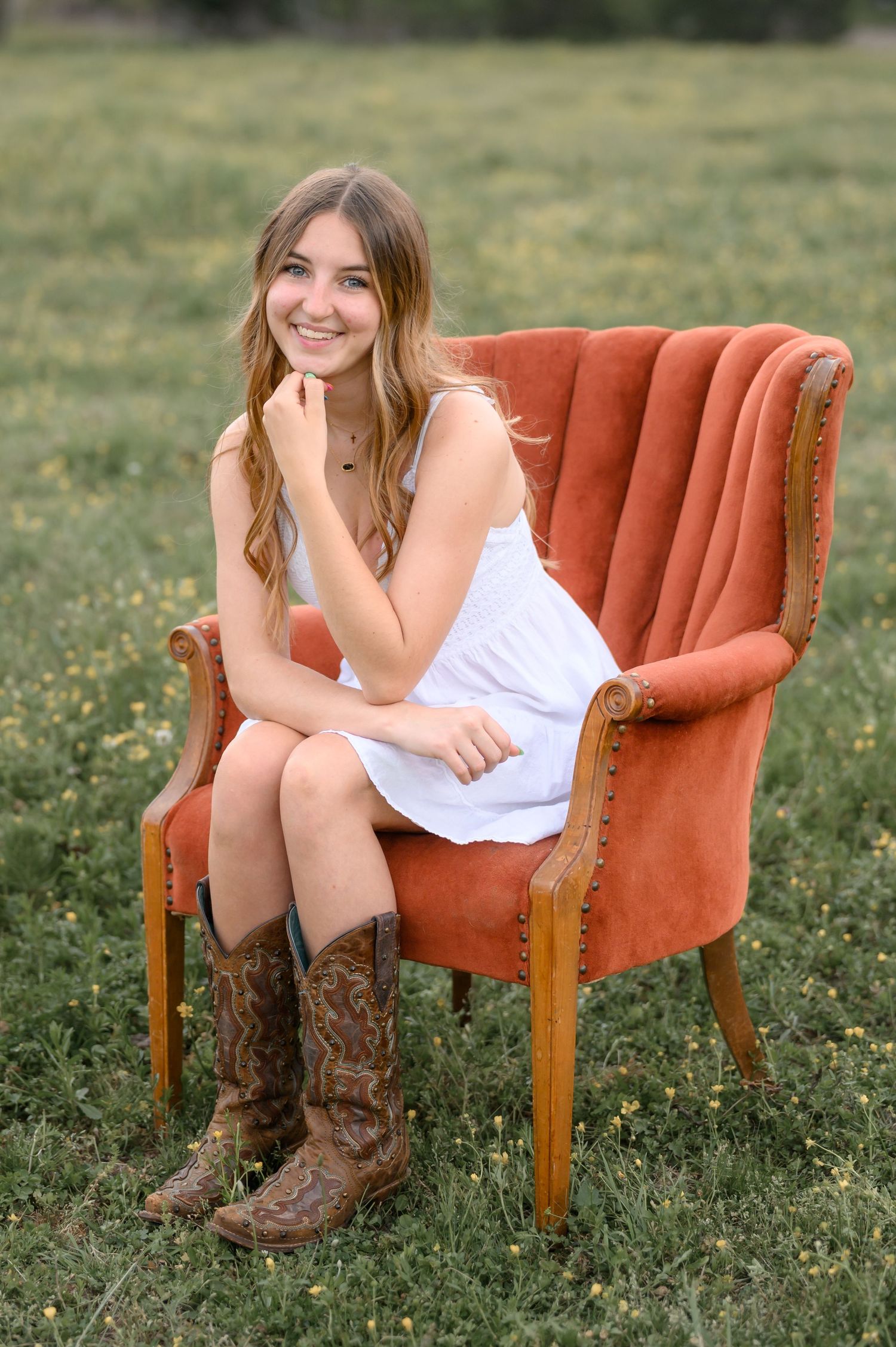 Smiling teenage girl in white dress and cowboy boots sitting in a rustic red velvet chair in a green field.