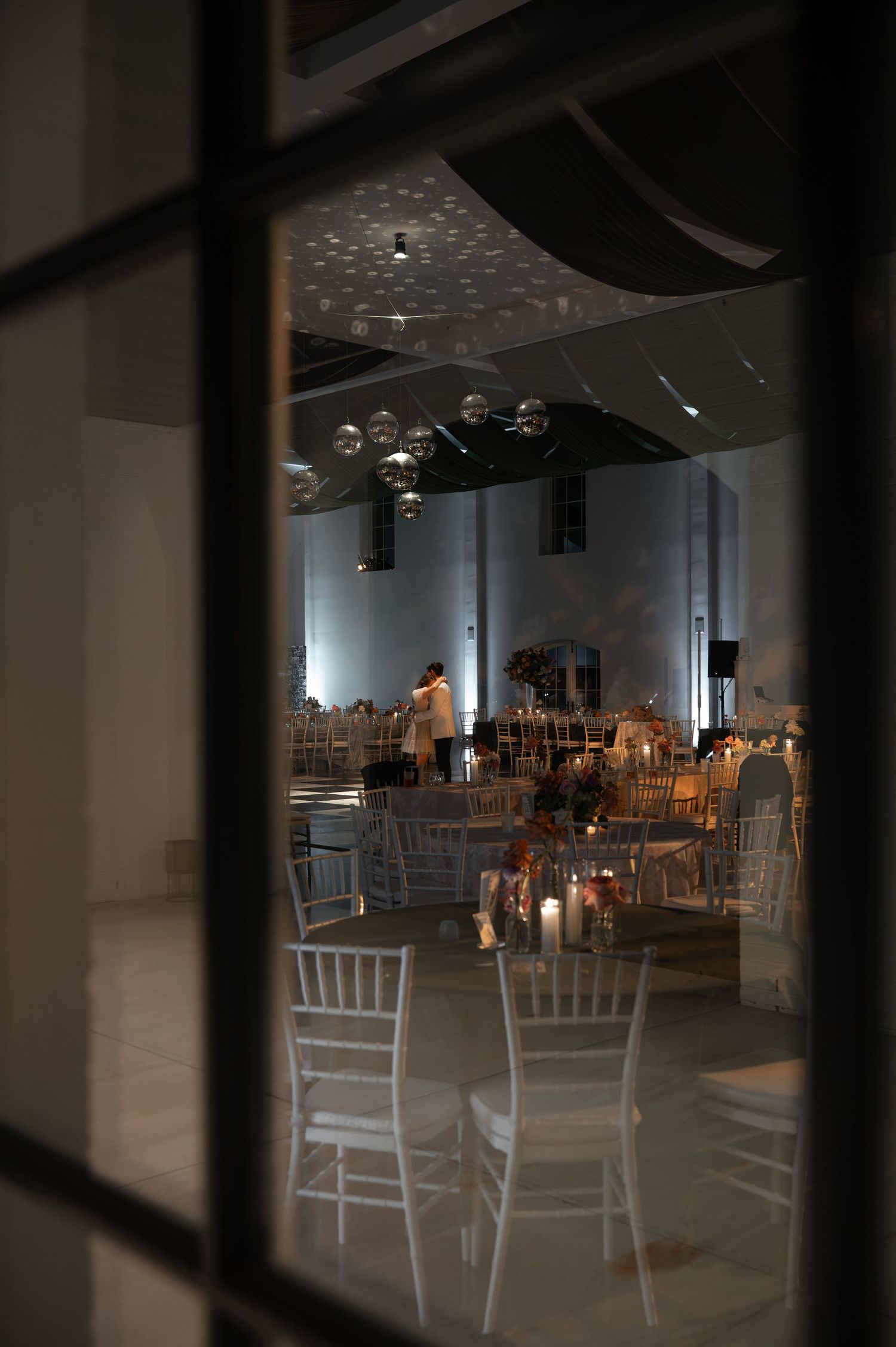 Elegant wedding reception venue viewed through window with white chiavari chairs, candles, and hanging bubble decorations.