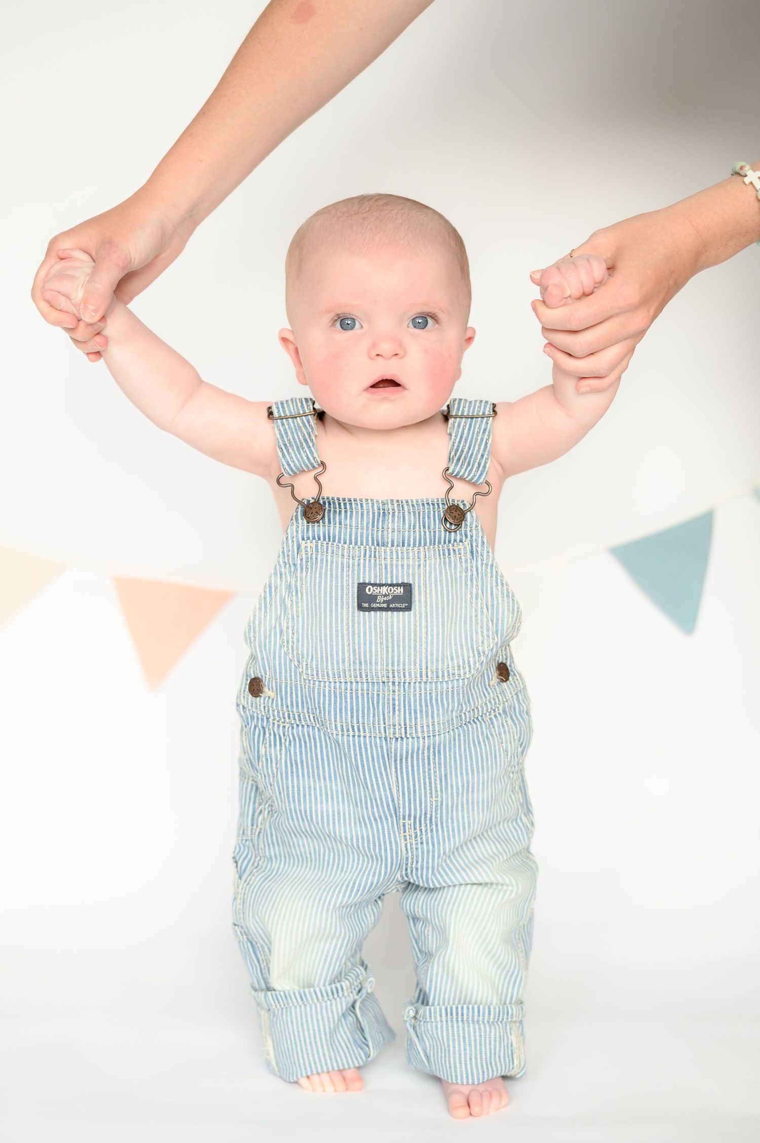 Adult hands helping blue-eyed baby in striped OshKosh overalls stand upright against white background with pennants.