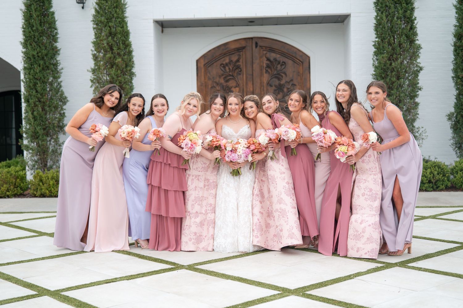 Bride with bridesmaids in mix of pink and mauve gowns holding floral bouquets.