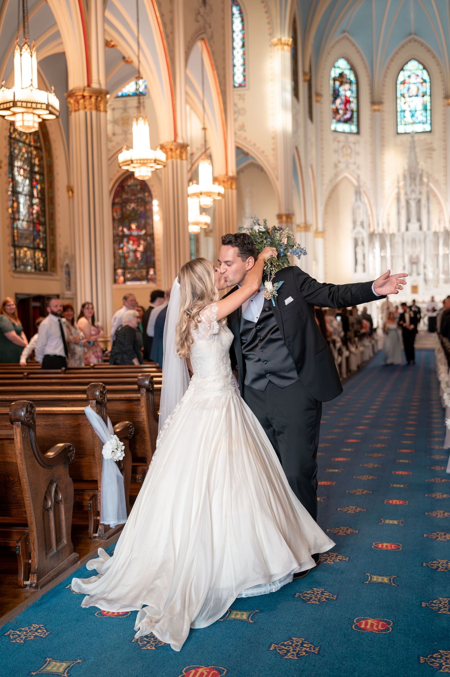 Newlyweds share kiss in grand cathedral aisle with blue carpet and stained glass.