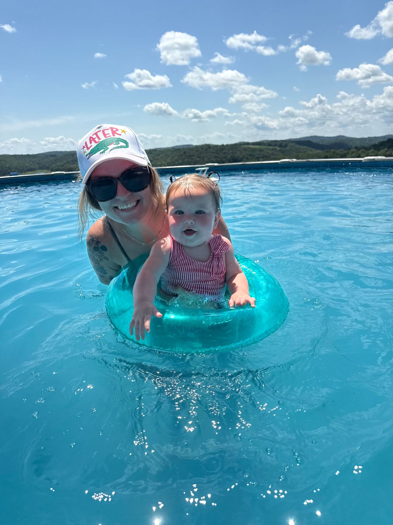 Woman in white cap and sunglasses helps smiling baby in teal pool float in bright blue swimming pool.