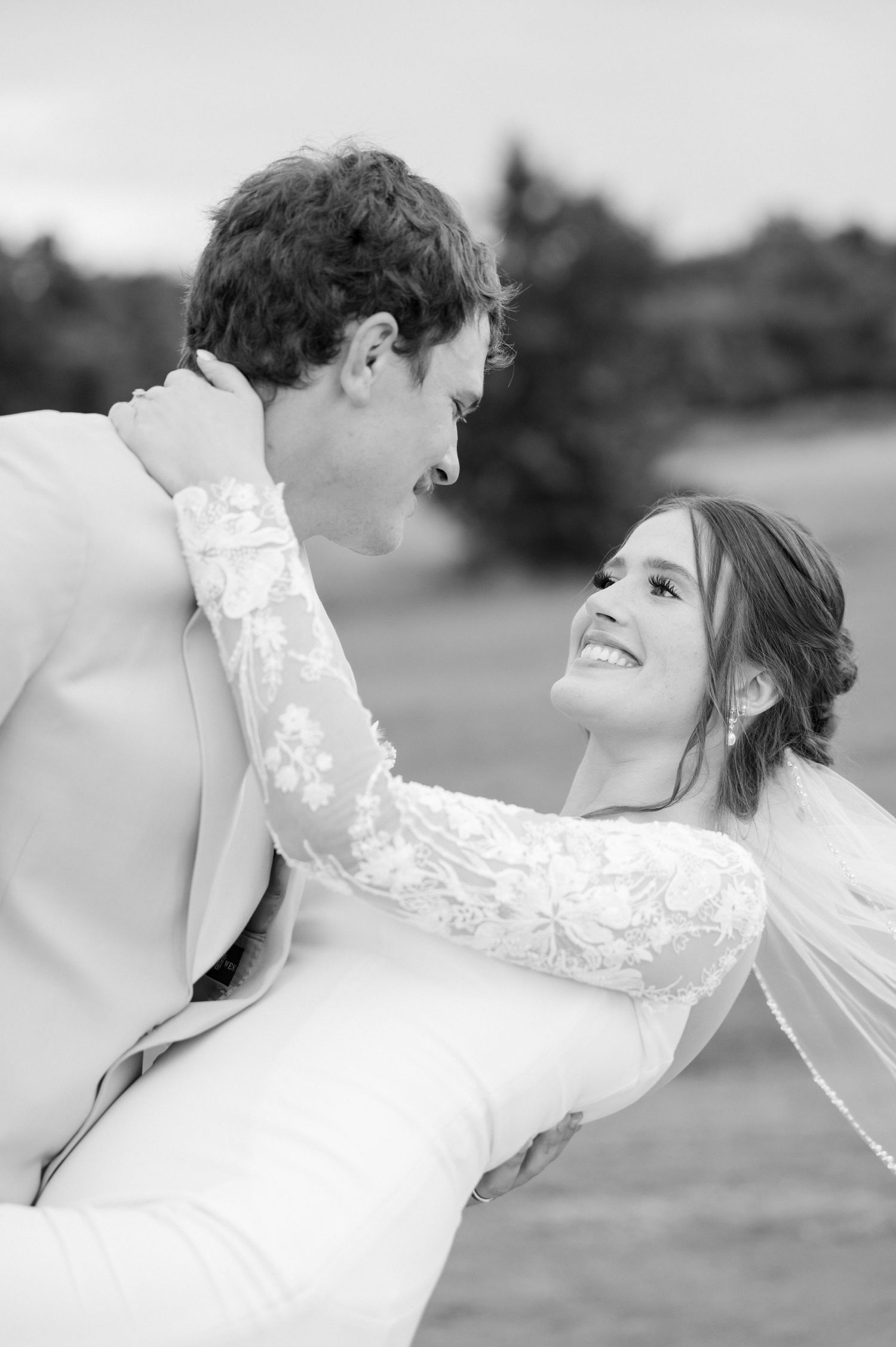 Black and white photo of a groom lifting his smiling bride in a lace wedding dress outdoors.