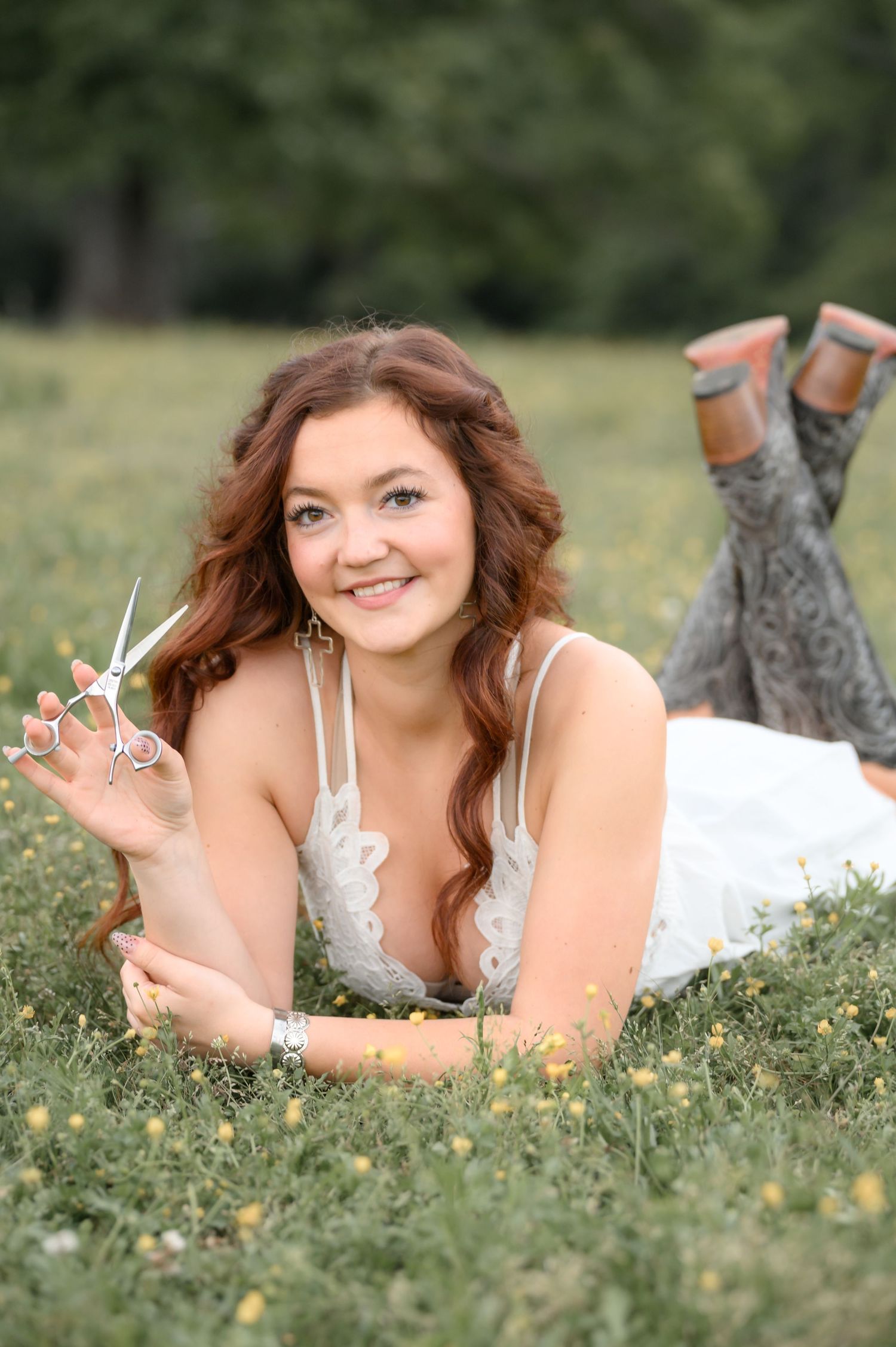 Smiling redhead woman in white dress lying in grass holding hair scissors, wearing cowboy boots.