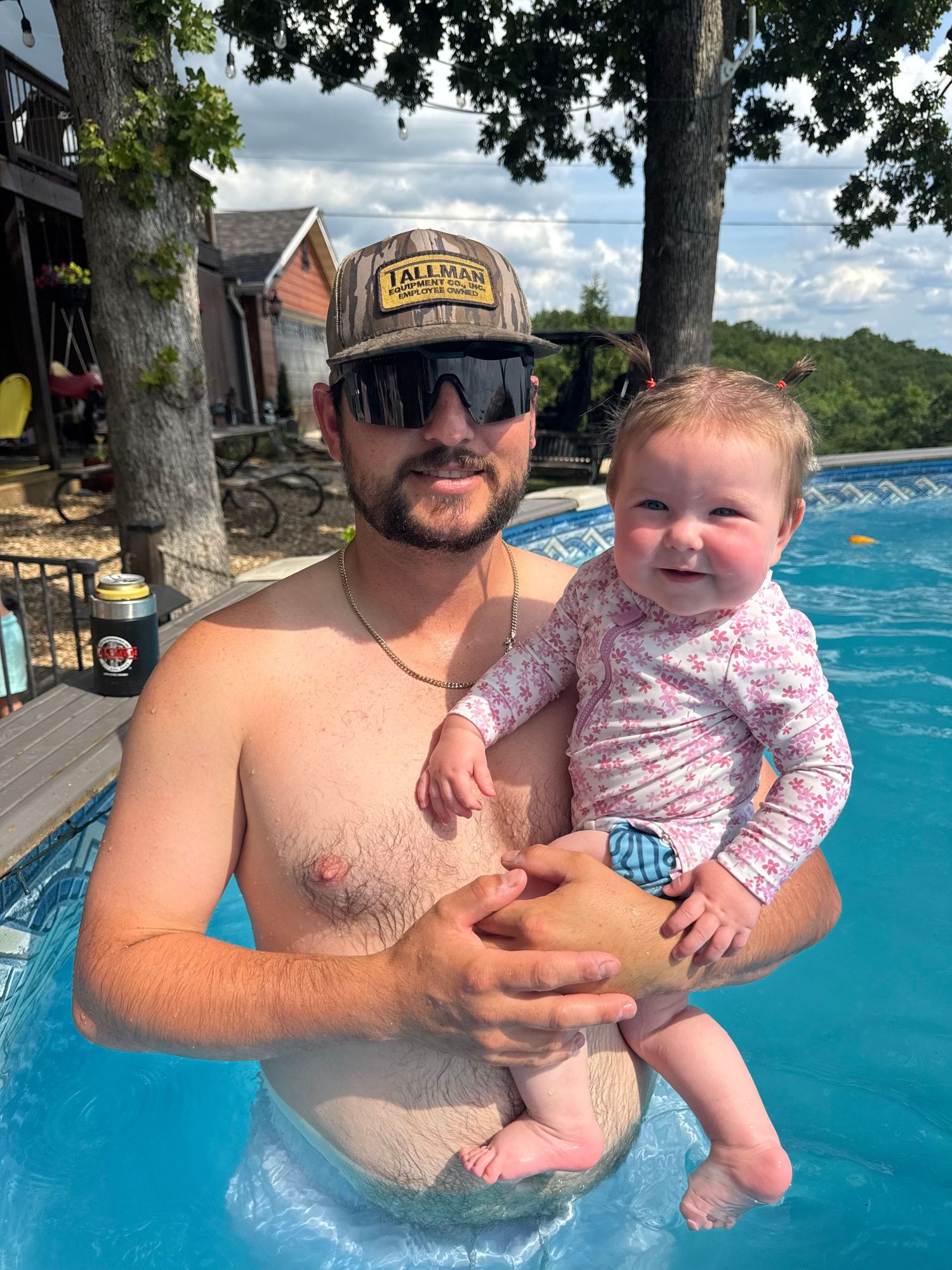 A shirtless man wearing sunglasses and a cap holds a smiling baby girl in a backyard swimming pool.
