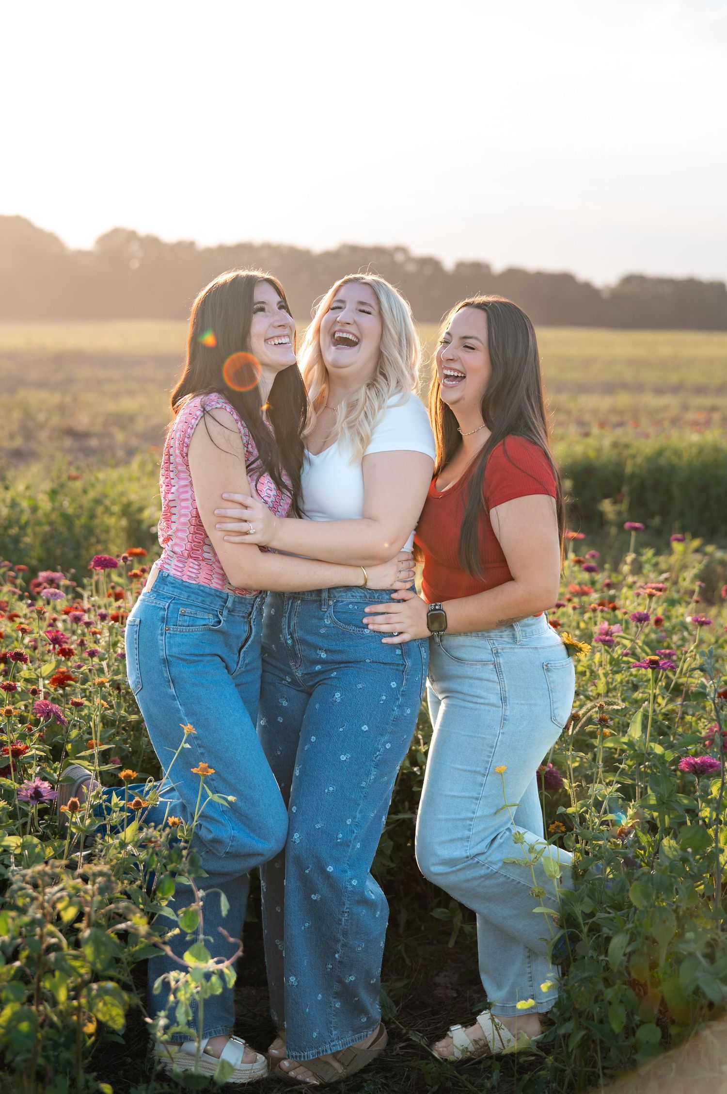 Three young women laughing together in a vibrant flower field during golden hour sunset.
