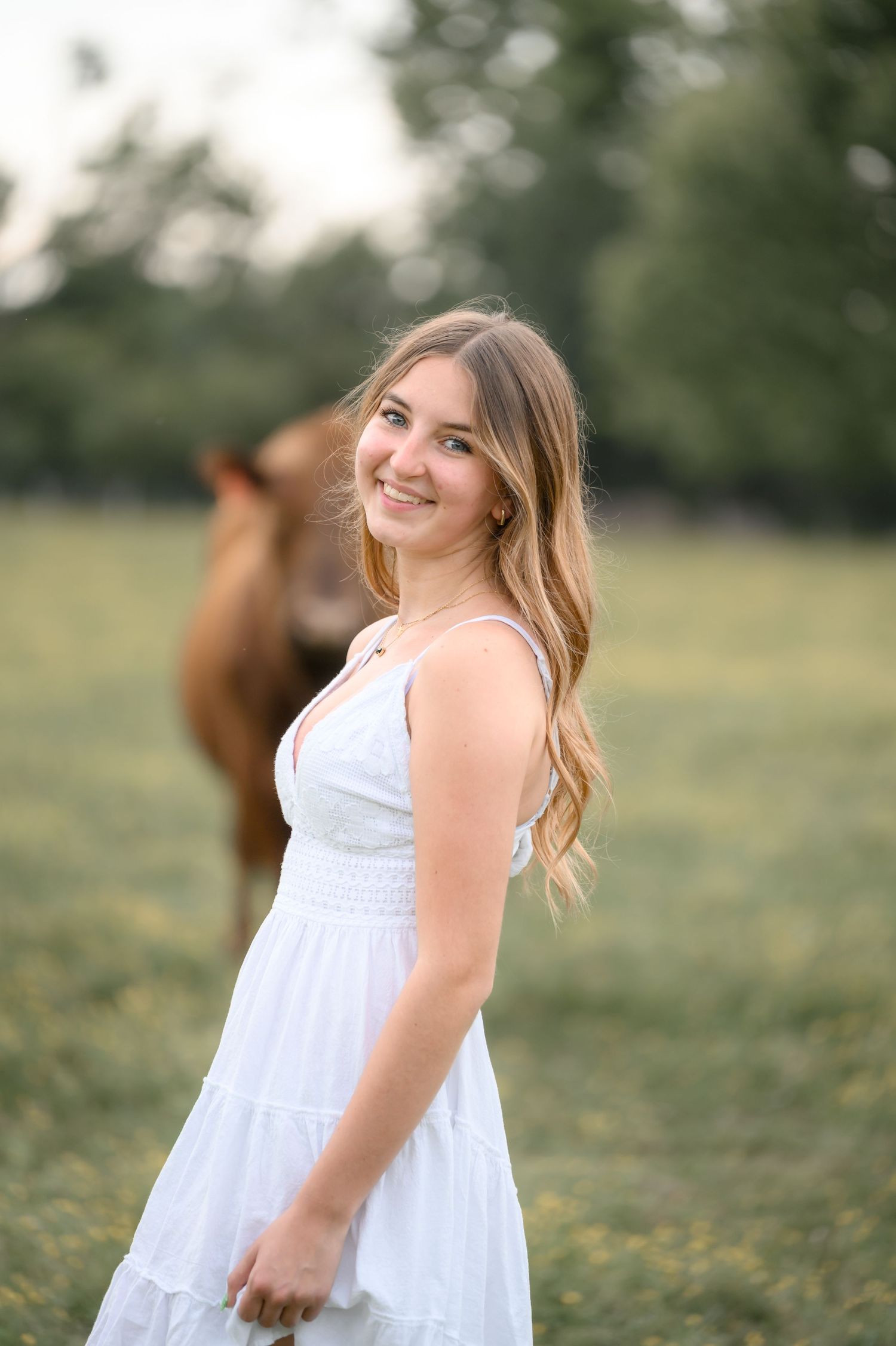 Smiling young woman in white sundress poses in golden field with a horse softly blurred behind her.