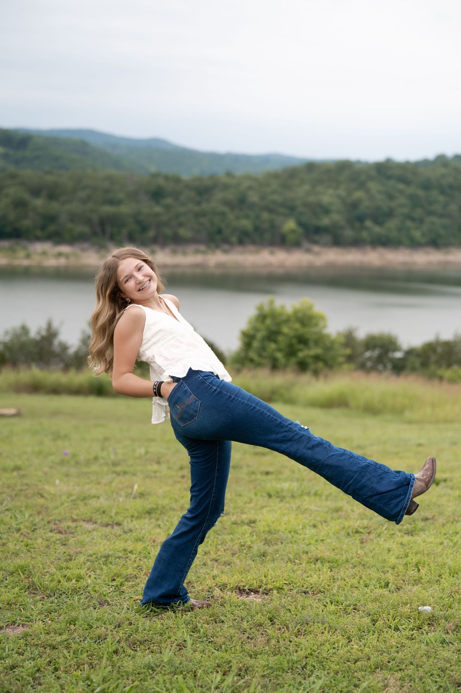 Teenage girl in white top and flare jeans playfully posing on green grass near a lake.