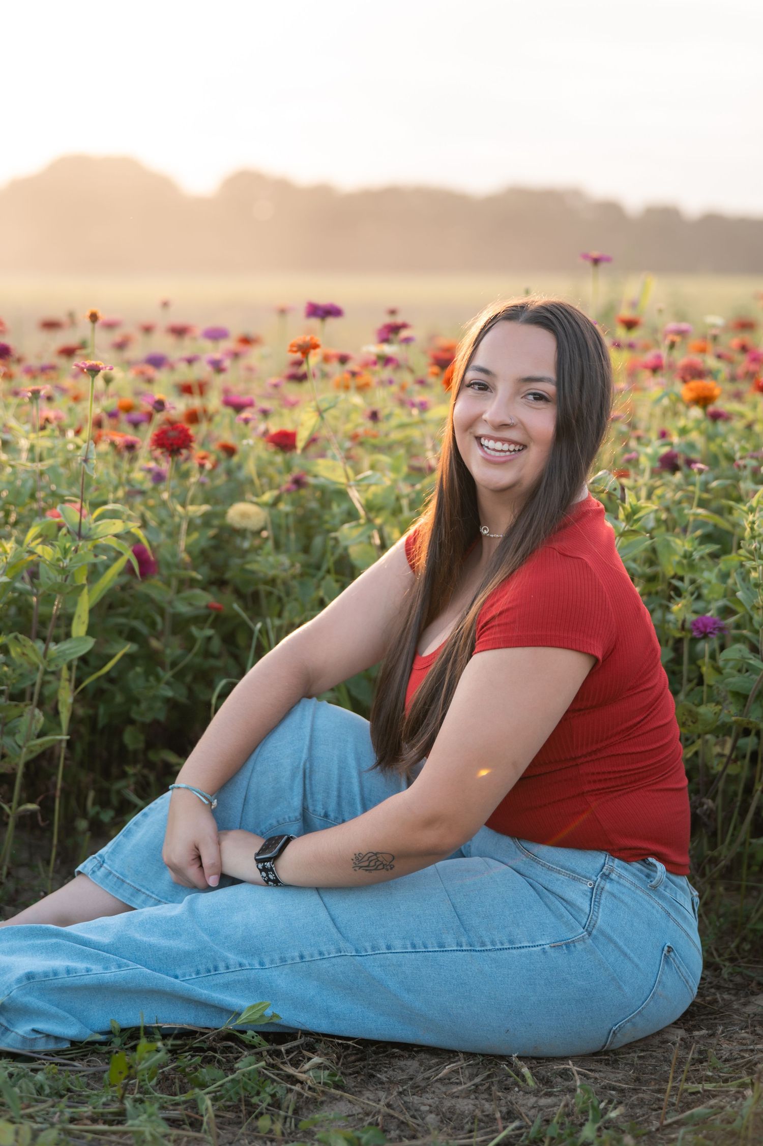 Smiling young woman in red shirt and jeans sitting in a vibrant zinnia flower field at sunset.
