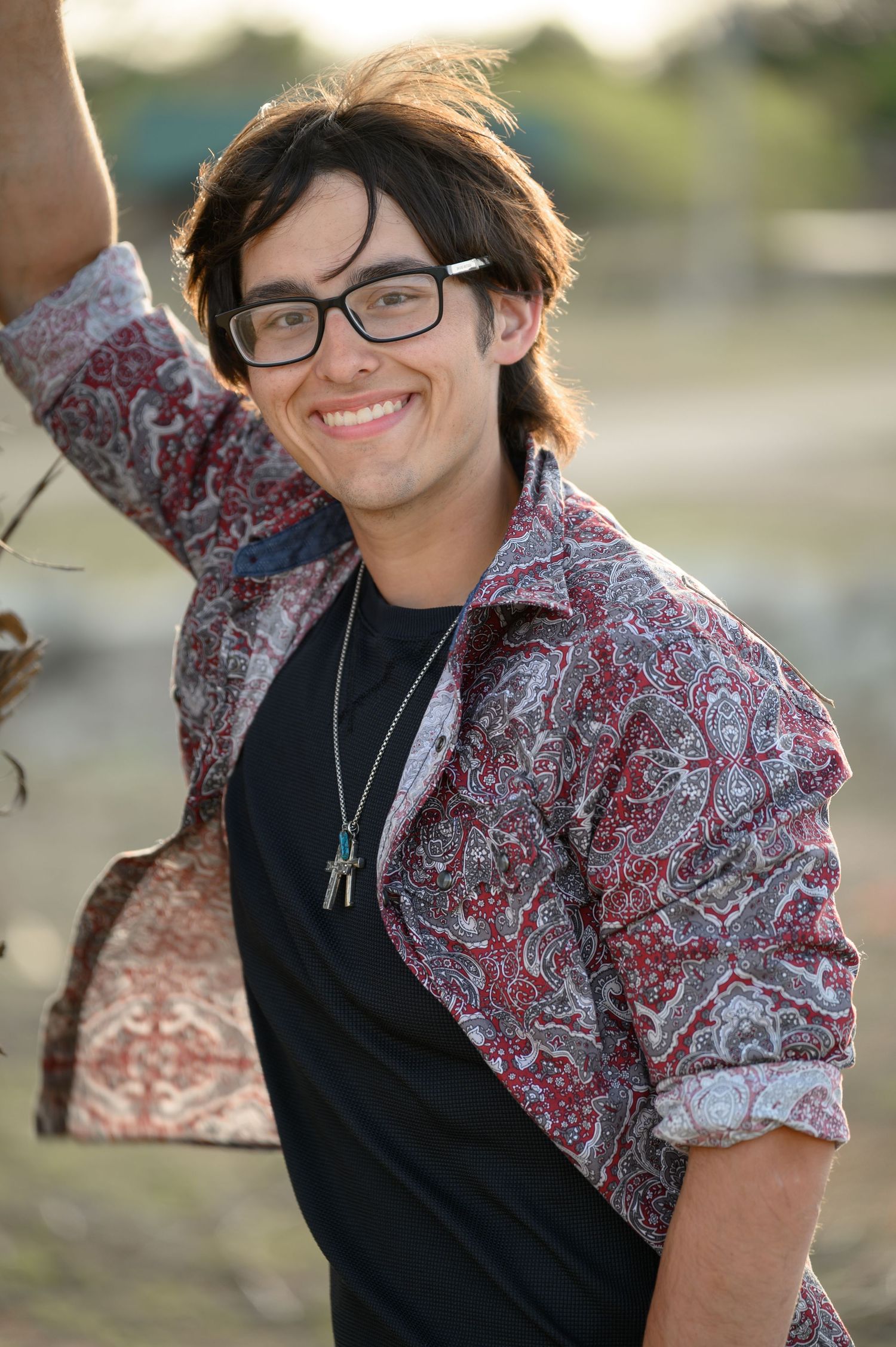 Smiling teenage boy with glasses and necklace leaning against a tree in a red paisley shirt outdoors.