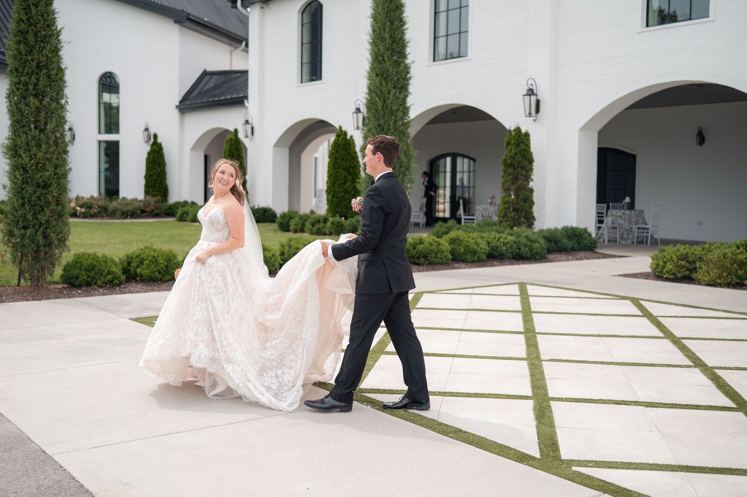 Bride in blush lace ballgown walks with groom in black tuxedo outside stunning white estate venue.