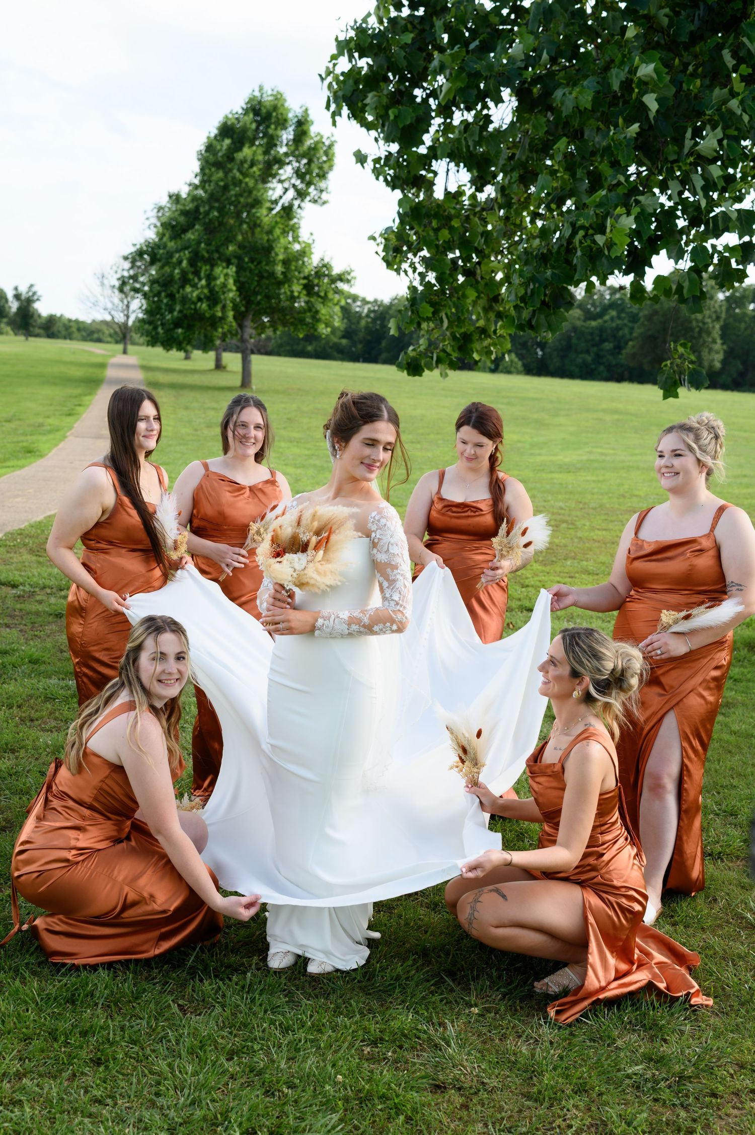 Bridesmaids in rust satin dresses help bride display her white wedding gown train on green lawn.