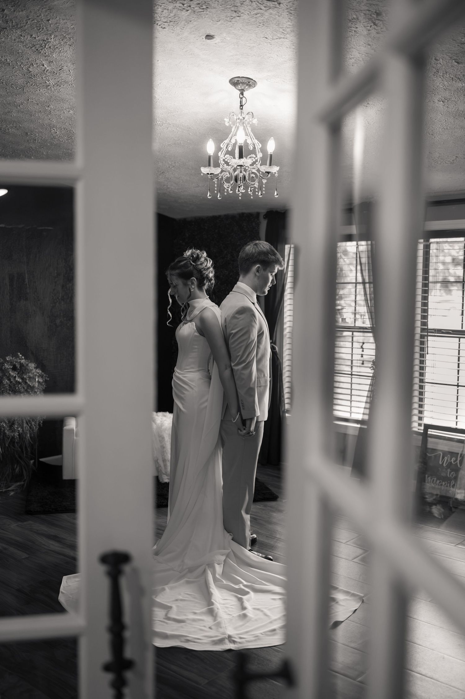 Black and white photo of bride and groom standing back-to-back in elegant room with chandelier, viewed through French doors.