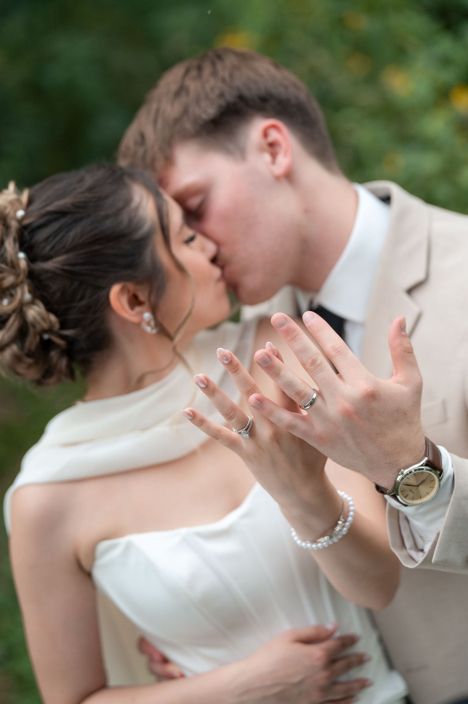 Newlyweds kissing while displaying wedding rings, bride in white dress, groom in beige suit, outdoor ceremony.