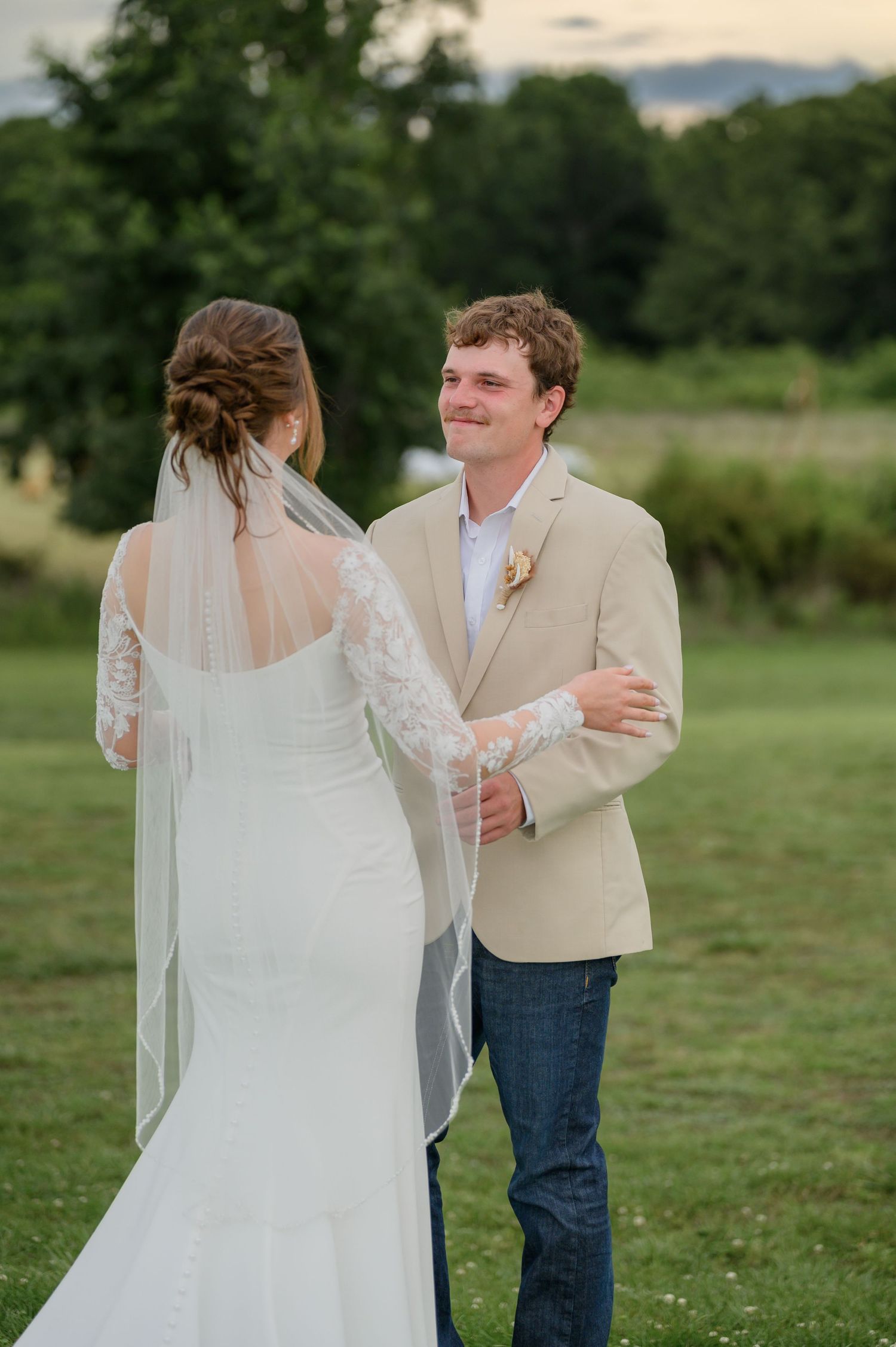 Smiling groom in tan blazer and jeans turns to see his bride during first look.