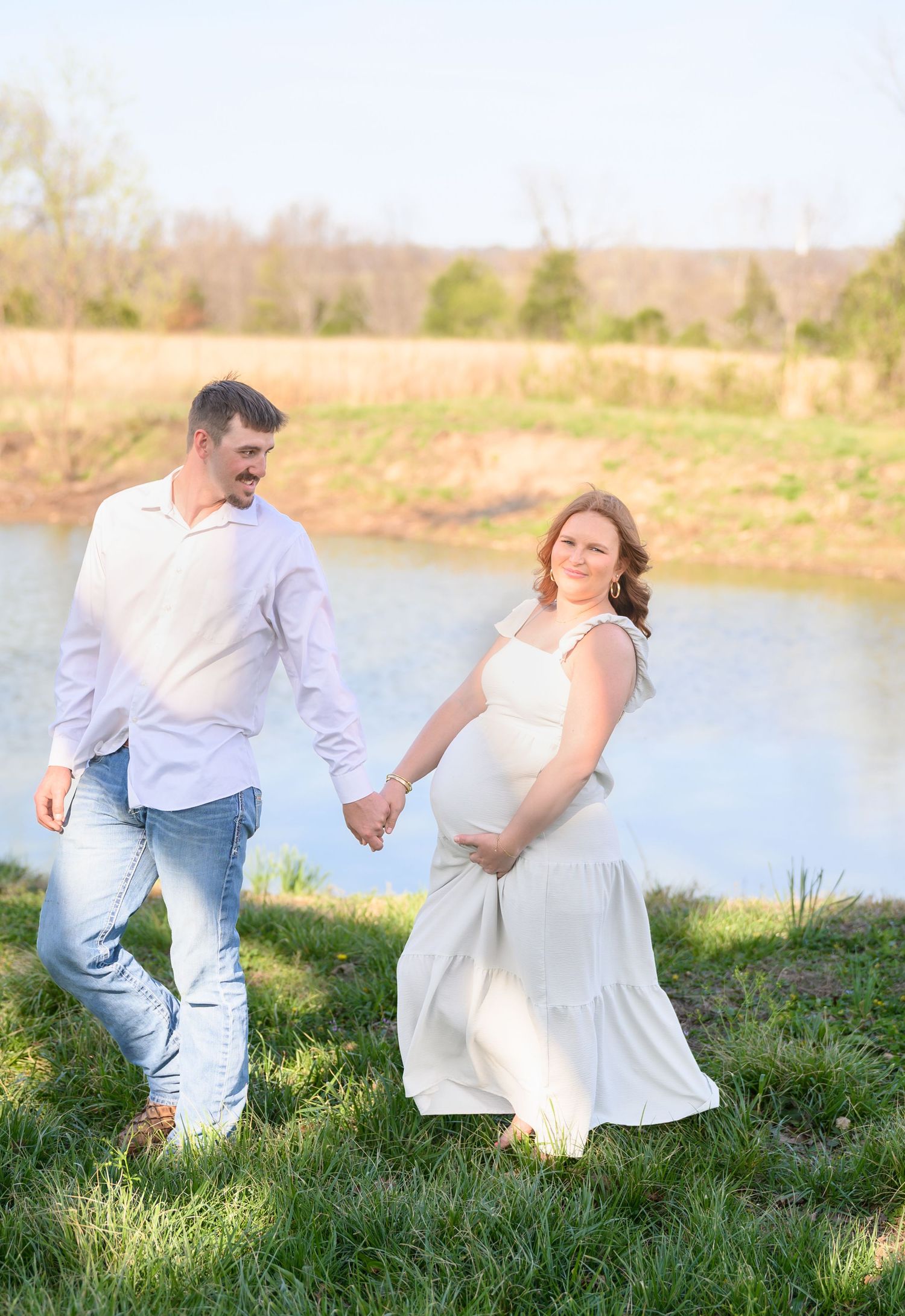 Pregnant couple holding hands walking near a pond in golden hour light.