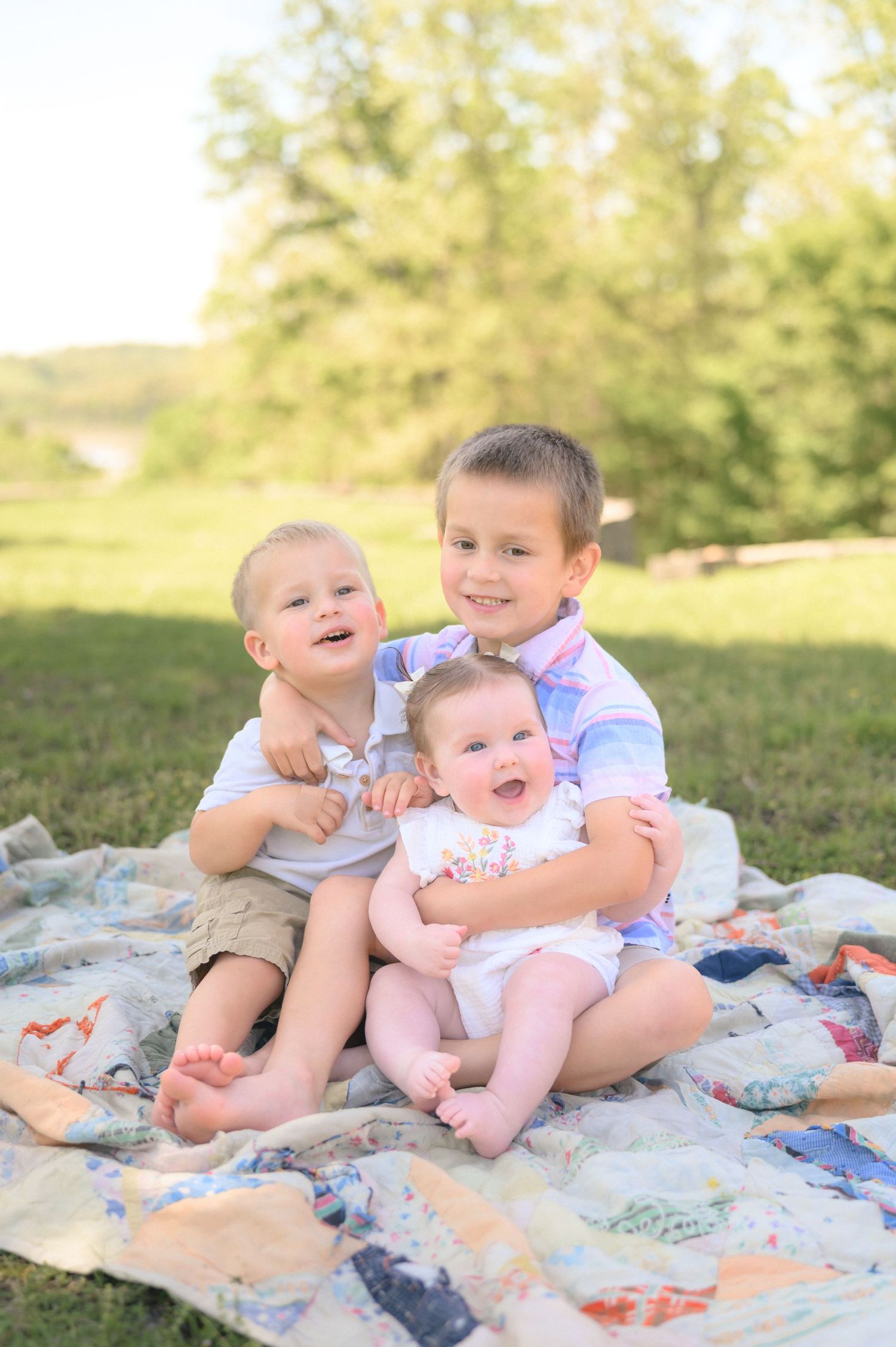 Three young siblings sitting on a quilt blanket outdoors, smiling and hugging in a sunny green park setting.