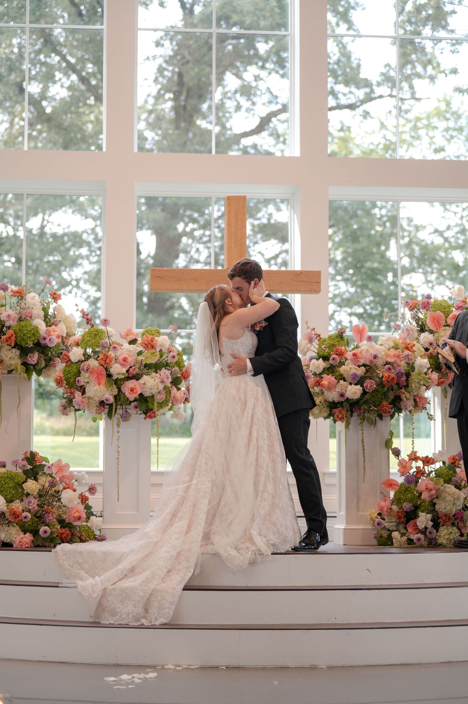Bride and groom kiss at floral-adorned altar with wooden cross backdrop.