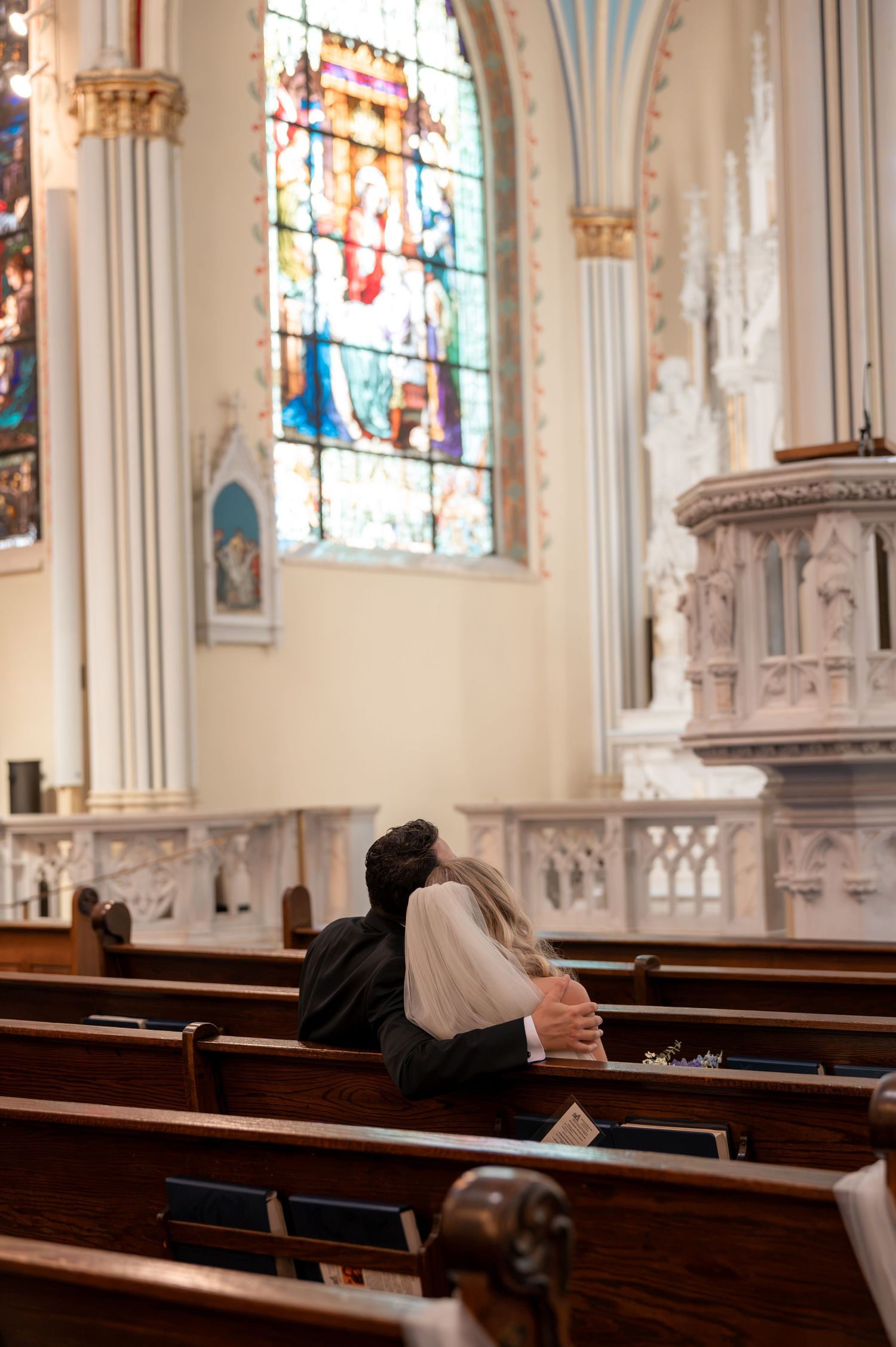 Bride and groom sit closely together in church pew beneath beautiful stained glass window.