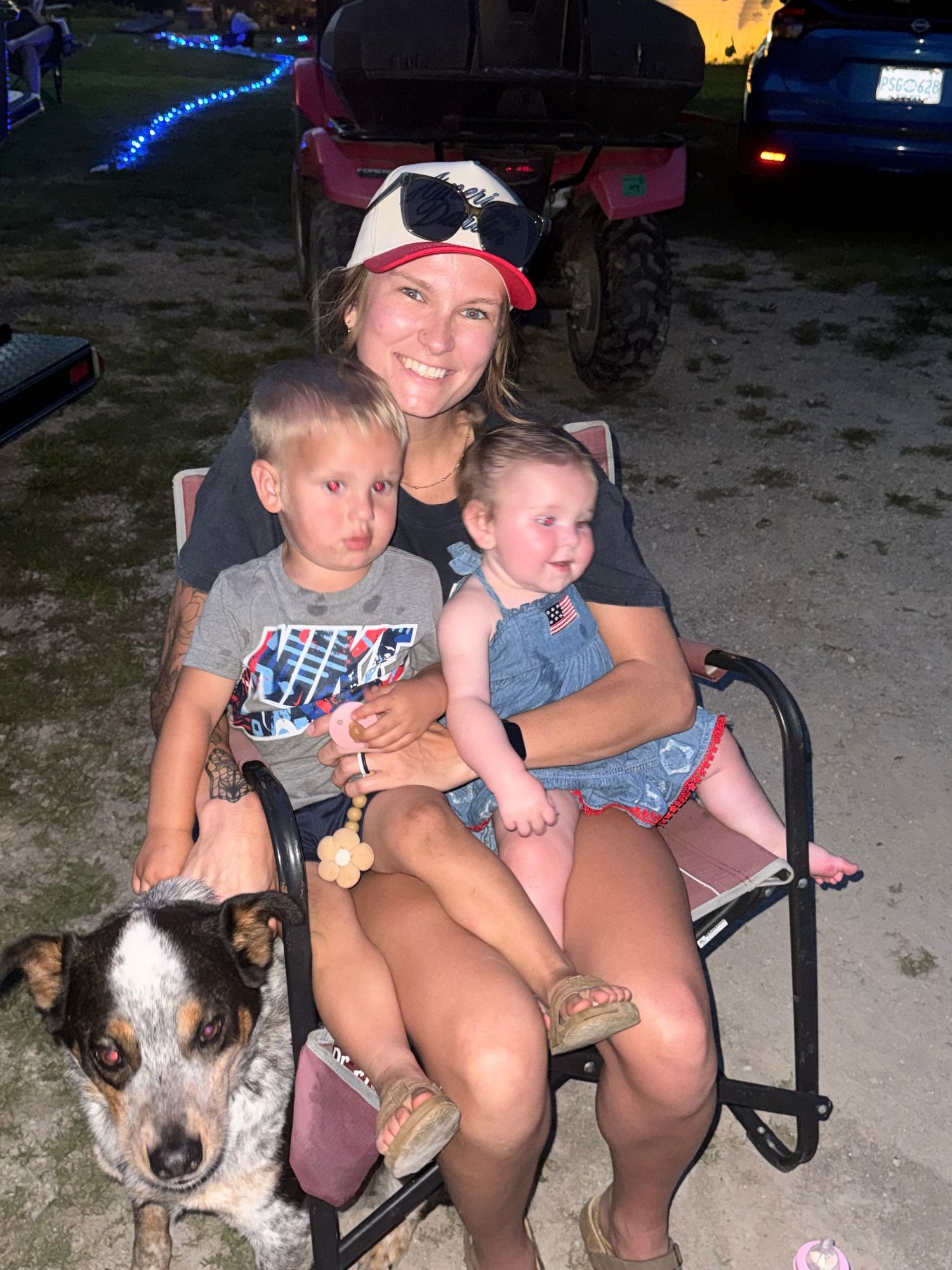 Woman in baseball cap sits in folding chair outdoors with two young children on her lap and a blue heeler dog beside her.