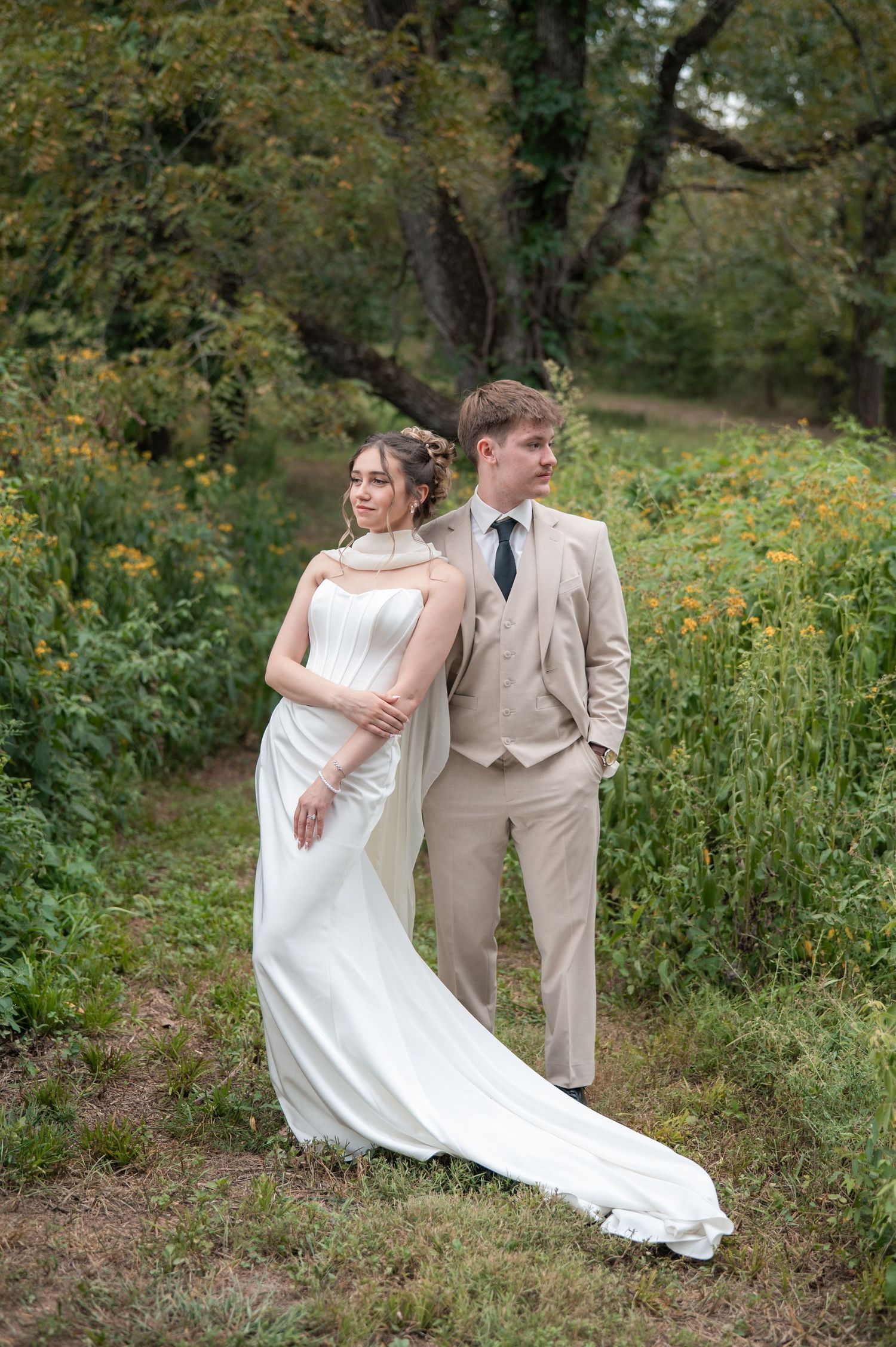 Bride in white gown and groom in beige suit pose together in a lush green outdoor setting.