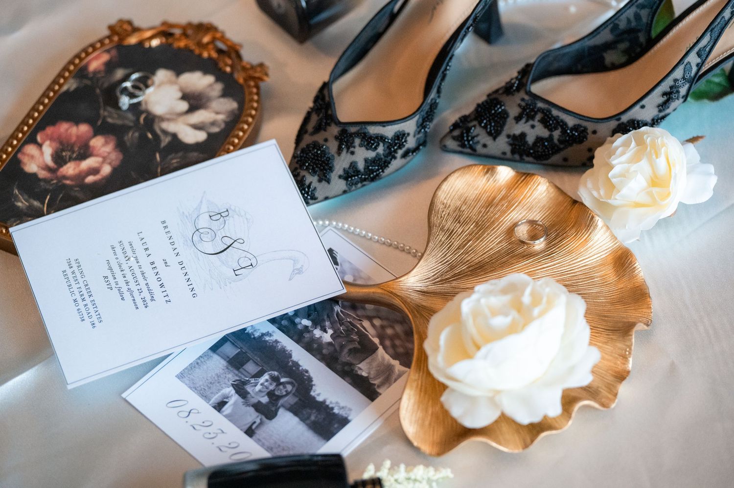 Elegant wedding flat lay with floral heels, invitation, gold shell dish, white roses, and vintage photos on a white surface.
