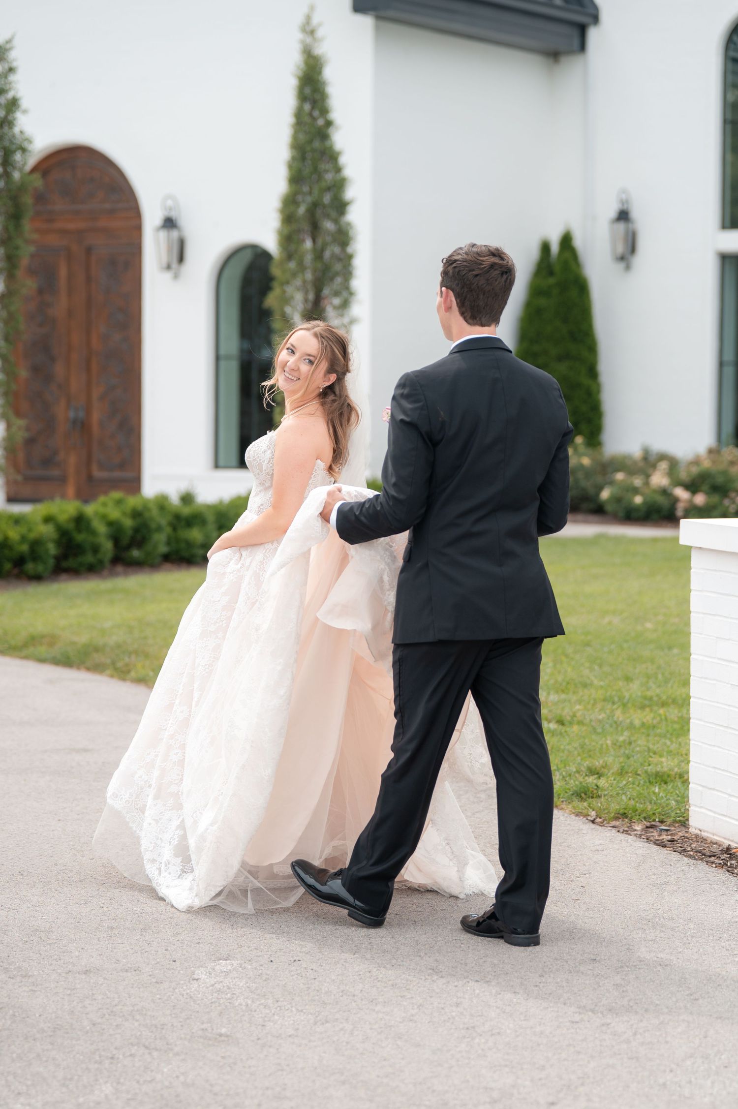 Smiling bride in lace ballgown glances back while groom guides her toward white wedding venue doors.