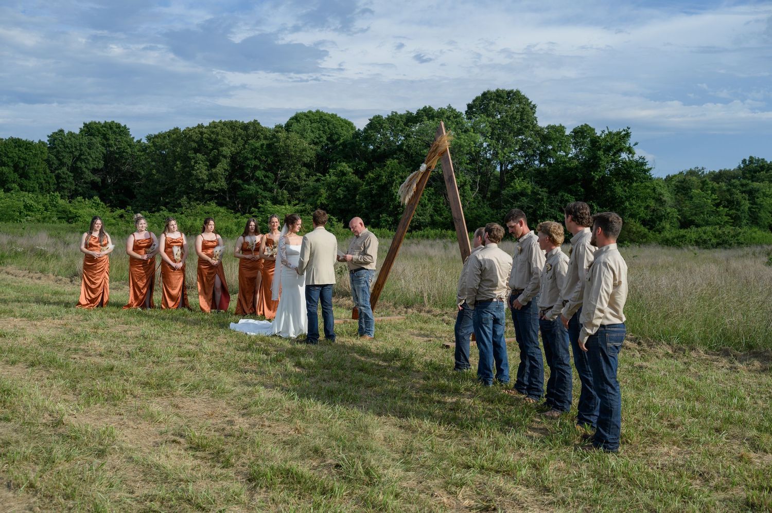 Outdoor rustic wedding ceremony with bridal party gathered under a wooden triangular arch in a meadow.