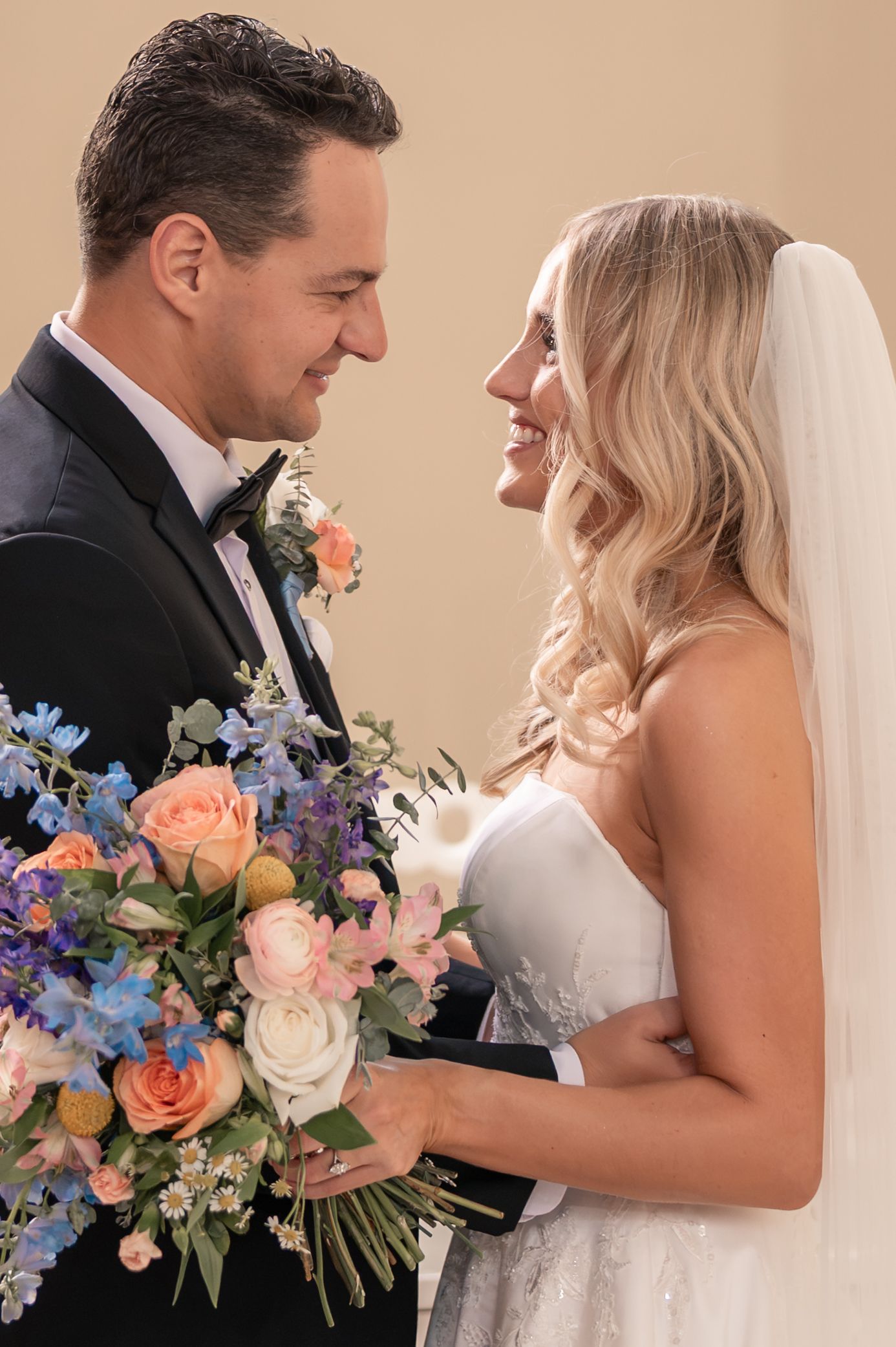 Smiling bride and groom gaze lovingly at each other while holding a colorful bridal bouquet of roses.