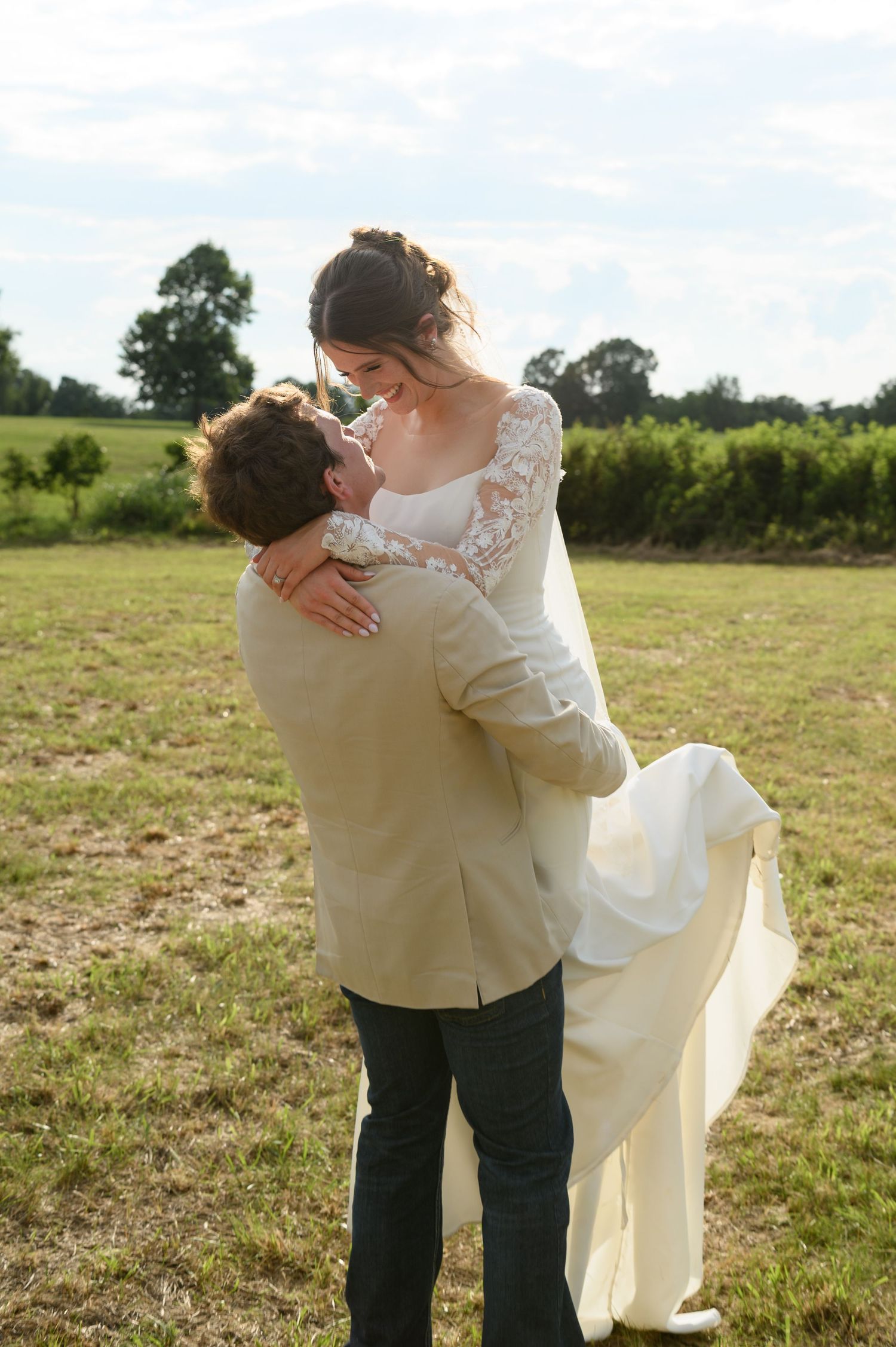 Groom lifting bride in white lace wedding dress outdoors in green field.