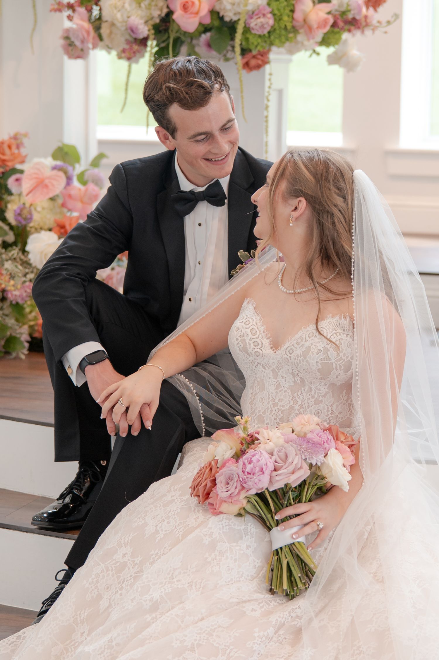 Bride and groom share a loving gaze while seated together surrounded by lush pink floral arrangements.