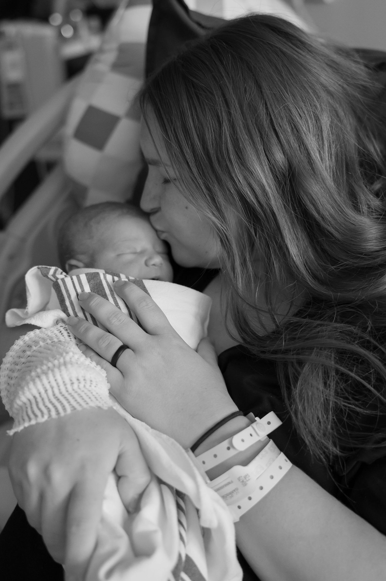 Black and white photo of a new mother kissing her swaddled newborn baby in a hospital room.